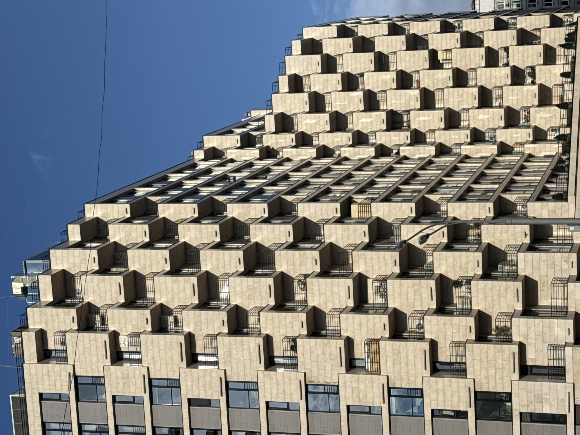 The image showcases a unique stepped facade of a building, likely an apartment complex, under a clear blue sky in Moscow, Russia. The building's design consists of receding tiers, each featuring box-like protrusions with what appear to be small balconies or recessed spaces. The color palette is dominated by beige and grey tones from the building's construction material and the reflection in the windows. The balconies have metal railings, adding a geometric element to the overall pattern. The rows of windows follow the stepped structure of the building, and at least one wire or cable is visible stretching across the sky in the upper left.