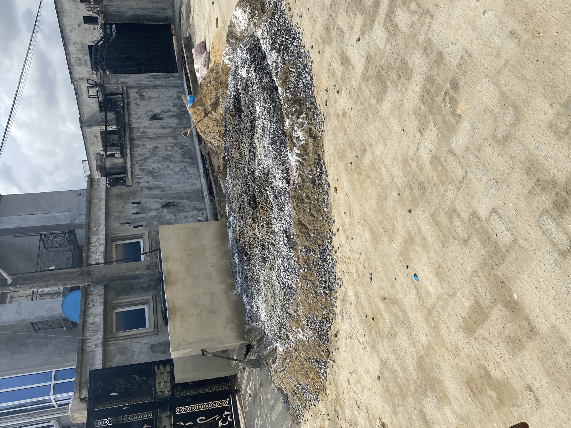 The image depicts a construction site in an urban environment in Unknown, Unknown. A pile of construction materials, likely gravel or crushed stone, is situated near a building undergoing renovation or construction. The building is multi-story and has windows, a gate, and some architectural details. The ground is paved, possibly concrete. There are no people visible in the frame.