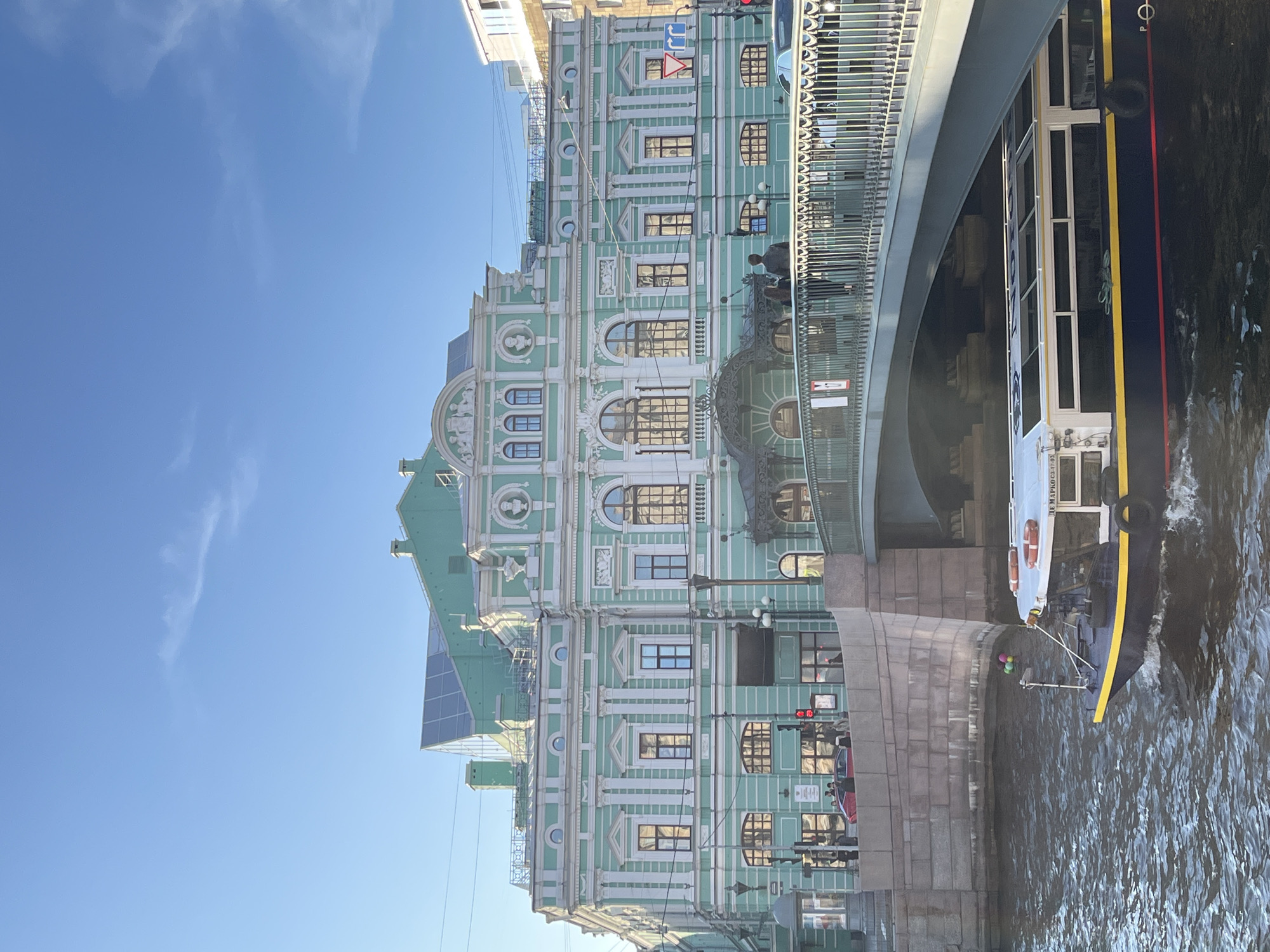 The image depicts a view in Saint Petersburg, Russia. A canal flows in the lower right corner, with a riverboat docked against a stone embankment. The boat is primarily dark blue with a yellow stripe and white window frames. A bridge spans the canal and boat.
To the left of the bridge, an ornate building with light green and white facade stands. The building has multiple stories with arched windows and decorative elements. A modern glass structure is attached to the building's upper portion. Several people stand on the bridge near the building. A traffic light is visible on the embankment. The sky above is clear and blue.