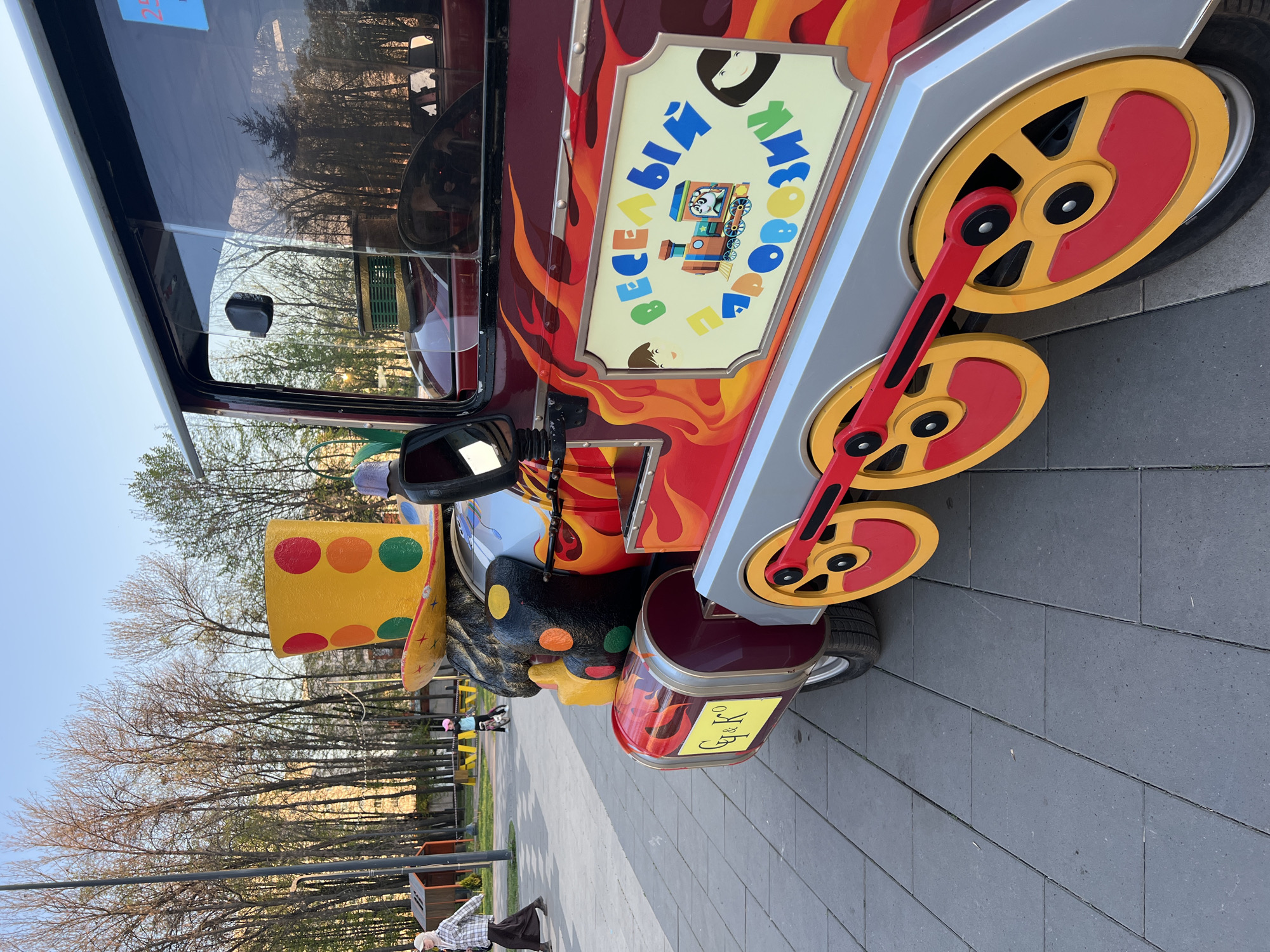 A vibrant, whimsically designed train-themed amusement ride stands stationary in a park setting, most likely in Ufa, Russia. The vehicle features bright red and yellow flame-like designs, oversized red and yellow wheels with connecting rods, and a clown-like figure with a yellow polka-dotted hat at the front. A sign with Cyrillic text and a cartoon train is attached to the side. The vehicle is parked on a gray brick pathway. In the background, leafless trees line the pathway and people are visible.