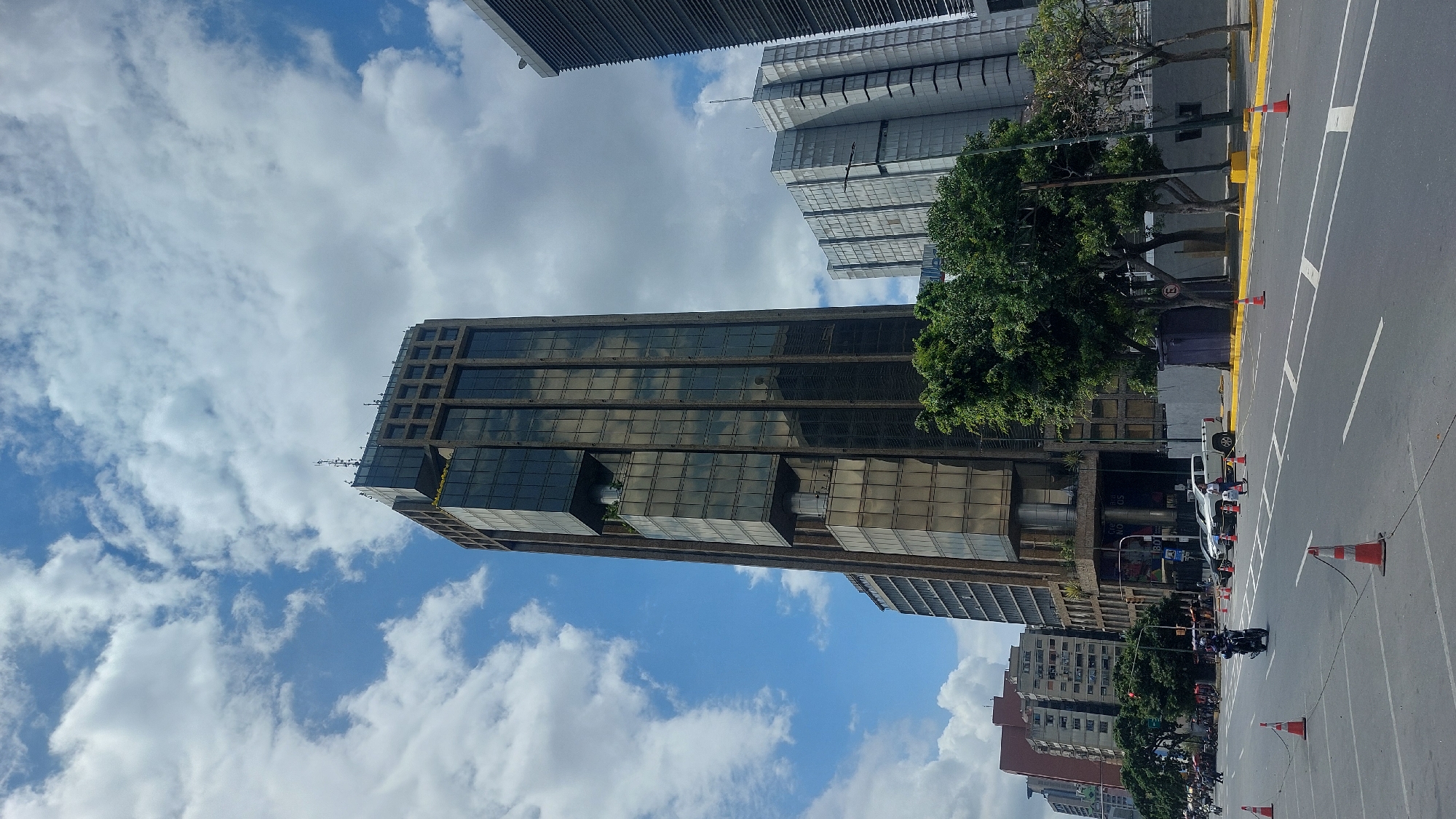 The image shows a view of a street in Caracas, Venezuela, with several buildings. The most prominent is a tall office building with a glass facade reflecting the sky. To the right, another modern building is seen with trees in front of it. The street has several lanes marked with white lines and orange traffic cones. There are cars parked on the side of the street and motorcycles. The sky is cloudy with blue patches visible.