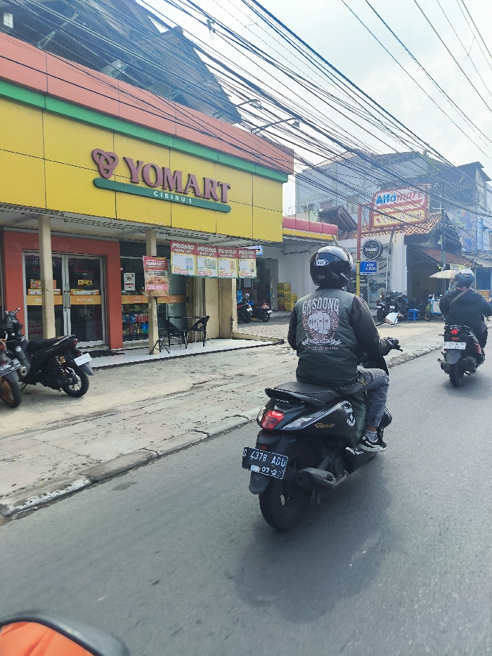 The image shows a street scene in Cileunyi, Indonesia (latitude: -6.933, longitude: 107.723). A man wearing a black and green jacket with the word "Gasdong" on the back is riding a black motorcycle, facing right. He has a black helmet on. His vehicle is a black Yamaha, with a license plate that reads "D 4378 ADU" and has a white sticker on the rear end. There are several other motorcycles parked on the sidewalk on the left, including a black and red one with a white license plate. The man is riding past a yellow building with the words "Yomart Cibeure 1" written on it. The building is a convenience store, and there are several signs in front of it. There is a small, red, and white building with the words "Alfamart" written on it, across the street to the right. The street is paved with asphalt and has a gray sidewalk on the left. The sky is blue with some white clouds. There are several power lines overhead. There is a black and red motorcycle parked on the sidewalk on the left. There is a black motorcycle in front of a building with the words "Alfamart" on it, on the right. There is a red and white motorcycle parked in front of a building with the words "Alfamart" on it, on the right. There is a person wearing a black jacket and a black helmet riding a black motorcycle in front of a building with the words "Alfamart" on it, on the right. The man is riding past the parked motorcycles, which are all facing forward. There is also a black and white motorcycle parked on the sidewalk, in front of the yellow building, on the left. There is a black and red motorcycle parked on the sidewalk, in front of the yellow building, on the left. There is a red and white motorcycle parked on the sidewalk, in front of the yellow building, on the left. The man is riding past all of these vehicles. The scene takes place during the day, and the weather is sunny.