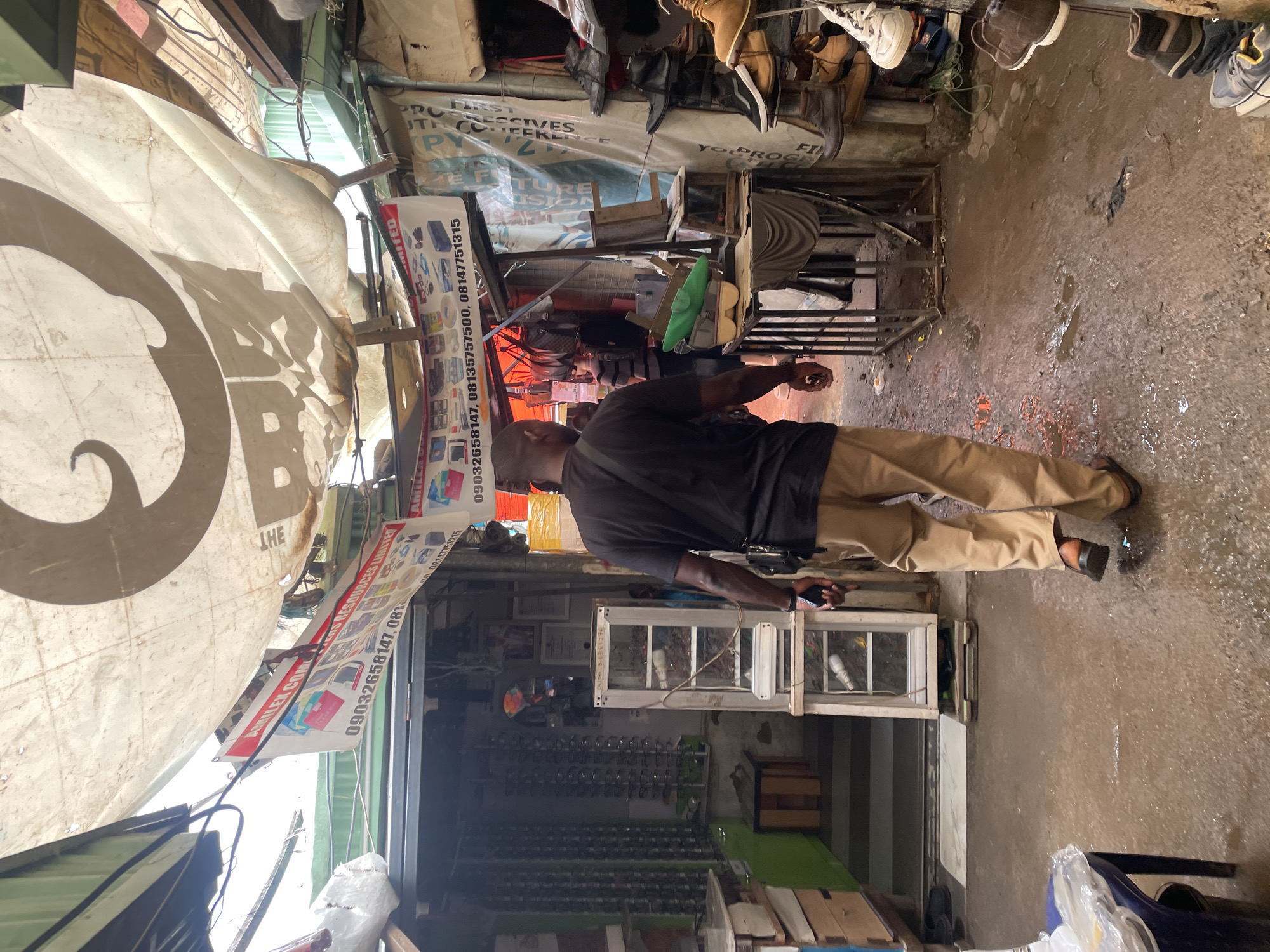 A man walks away from the camera down a narrow, outdoor market alleyway in Abuja, Nigeria. The ground is wet and muddy. The man is wearing a black t-shirt and tan pants. He has a black bag slung over his shoulder. In the background, there are market stalls with various goods displayed, including what appears to be an optician's shop with eyeglasses and frames on display. Tarps and awnings are strung overhead, providing shade and shelter. The scene suggests a busy, somewhat makeshift commercial environment.
