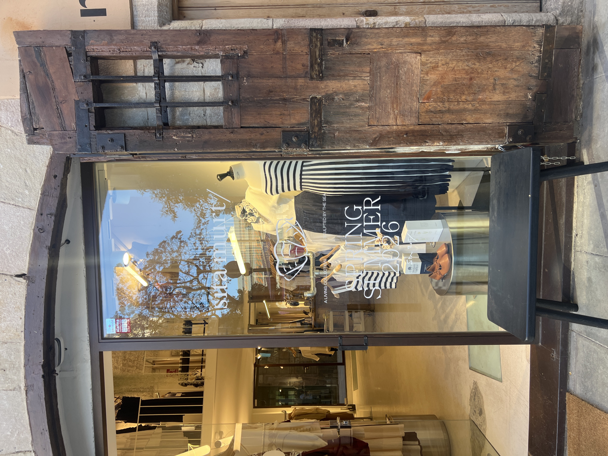 The image displays the entrance of a "sita murt/" clothing store, located in what appears to be a historic urban setting, specifically in Barcelona, Spain, given the architectural style and branding. The entrance consists of a modern glass double door storefront on the left, set within an older, rustic stone and dark wood archway. To the right of the glass doors is a weathered, dark brown wooden half-door or shutter, featuring black metal grates over an unglazed opening, secured by visible metal hinges and bolts. This wooden structure also has a large, rough-hewn wooden frame around it, indicating its age and traditional construction.
Inside the glass storefront, several mannequins display items from the "SPRING SUMMER 2026" collection. One prominent mannequin on the right wears a black and white striped strapless top paired with a long, vertically striped black and white skirt. Another mannequin to its left wears a light cream-colored knit top and a dark skirt. Text visible on the glass reads "sita murt/", "A LIVING COLLECTION," and "SCULPTED BY THE SEA." Below the mannequins, on a cylindrical silver pedestal, a pair of light brown leather shoes and several light-colored boxed items are displayed. Further inside the store, additional clothing racks with garments are visible, along with warm interior lighting and a clean, light-colored floor. A security sticker for "VERISURE Alarma" is affixed to the upper right pane of the glass door.
Reflections on the glass storefront show the clear sky and the branches of a tree, suggesting an outdoor setting during daylight hours. A black, rectangular wooden bench with four legs is positioned directly in front of the glass doors on the grey stone pavement. A brown doormat is partially visible on the far left of the foreground. The upper portion of the building above the archway is constructed from light-colored stone blocks.