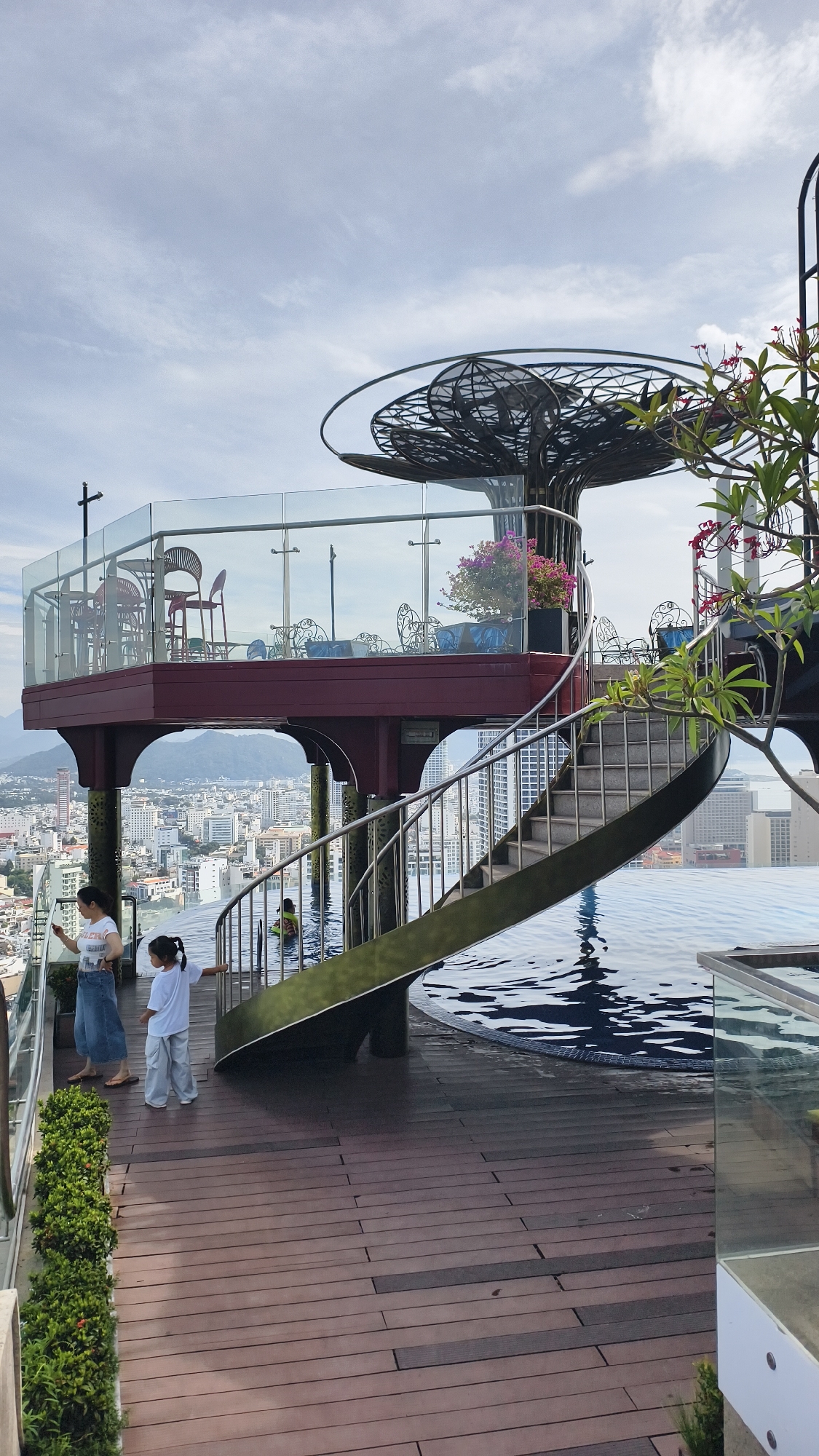 This is a vertical, eye-level, medium shot of the rooftop area of a building in Nha Trang, Vietnam. In the foreground, a wooden deck extends into the frame from the bottom left, with a row of green bushes lining the left edge. Two young females stand on the deck, one in a white t-shirt and blue jeans, holding a phone, and the other in a white t-shirt and light blue pants, facing away from the camera. Adjacent to the deck, a pool of dark blue water with ripples is visible on the right. A silver spiral staircase with a green accent curve ascends from the pool area to an upper level. The upper level features a maroon platform with a glass railing, furnished with chairs and tables, and a large, stylized metal tree sculpture. In the background, the city of Nha Trang stretches out, with numerous tall buildings visible, leading to a hazy horizon with mountains. The sky is overcast with scattered clouds.
