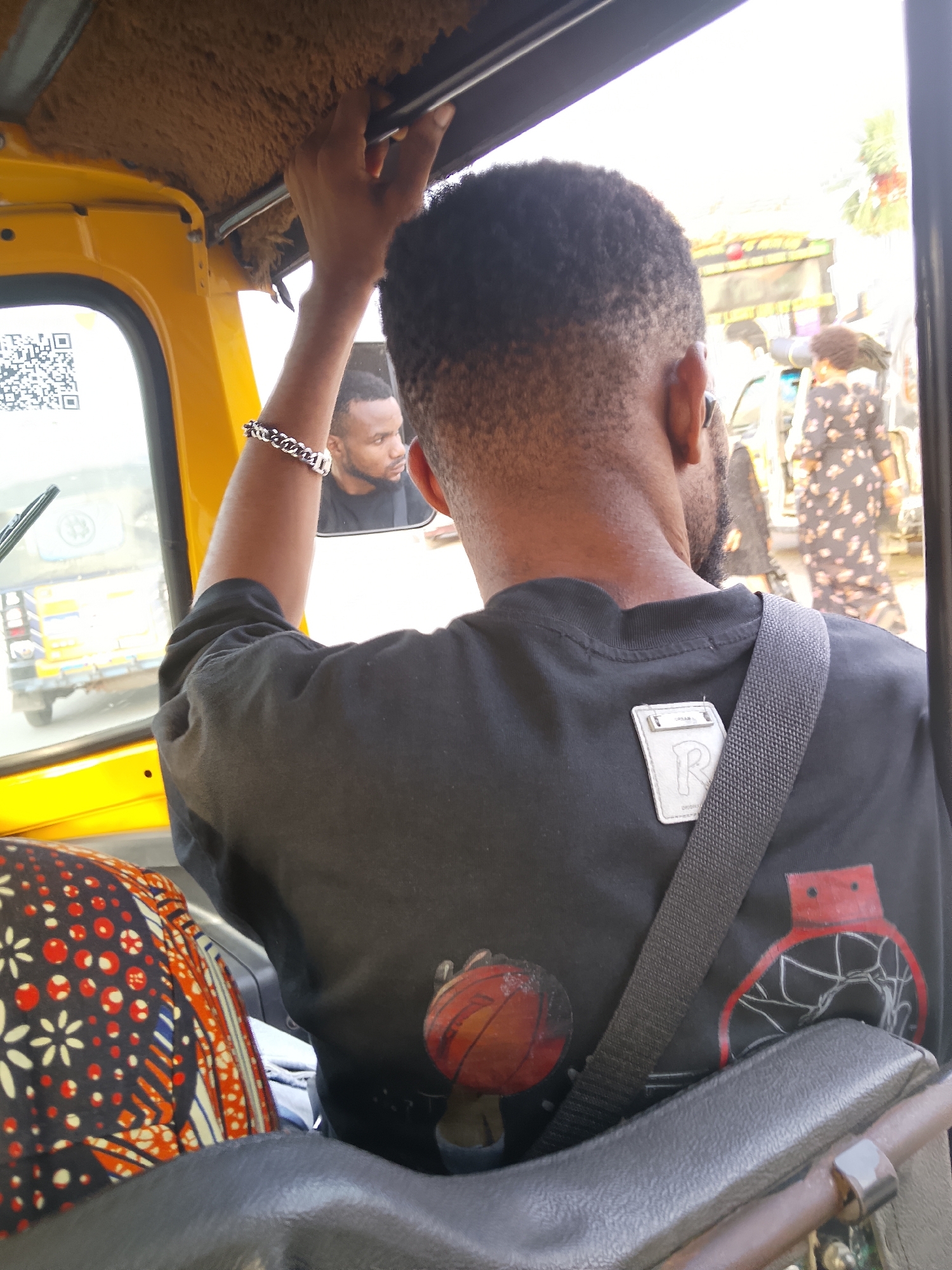 The image depicts a vibrant daytime street scene in Fegge, Nigeria, observed from inside a yellow Keke Napep (tricycle taxi). The primary subject is a dark-skinned man, seen from behind, occupying a passenger seat. He has short, dark, curly hair and a beard. He wears a black t-shirt adorned with a graphic of a basketball and a red basketball hoop on the lower back. A white patch featuring a prominent "R" and the word "ORIGINAL" beneath it is affixed to the upper back of his shirt, partially obscured by a black seatbelt strap. A silver chain bracelet is visible on his left wrist.
The man's face is reflected in the rearview mirror, showing him looking forward and slightly to his left with a neutral expression. His right hand is extended upwards, grasping a bar on the Keke's plush, brown-textured ceiling.
Through the Keke's window, bright daylight illuminates the surroundings. Another yellow Keke Napep is visible ahead, featuring a QR code sticker on its window. Blurred figures in the background, including a person in a patterned dress, suggest a bustling street with pedestrians and other vehicles. The scene captures an everyday moment of transportation in a Nigerian urban setting.