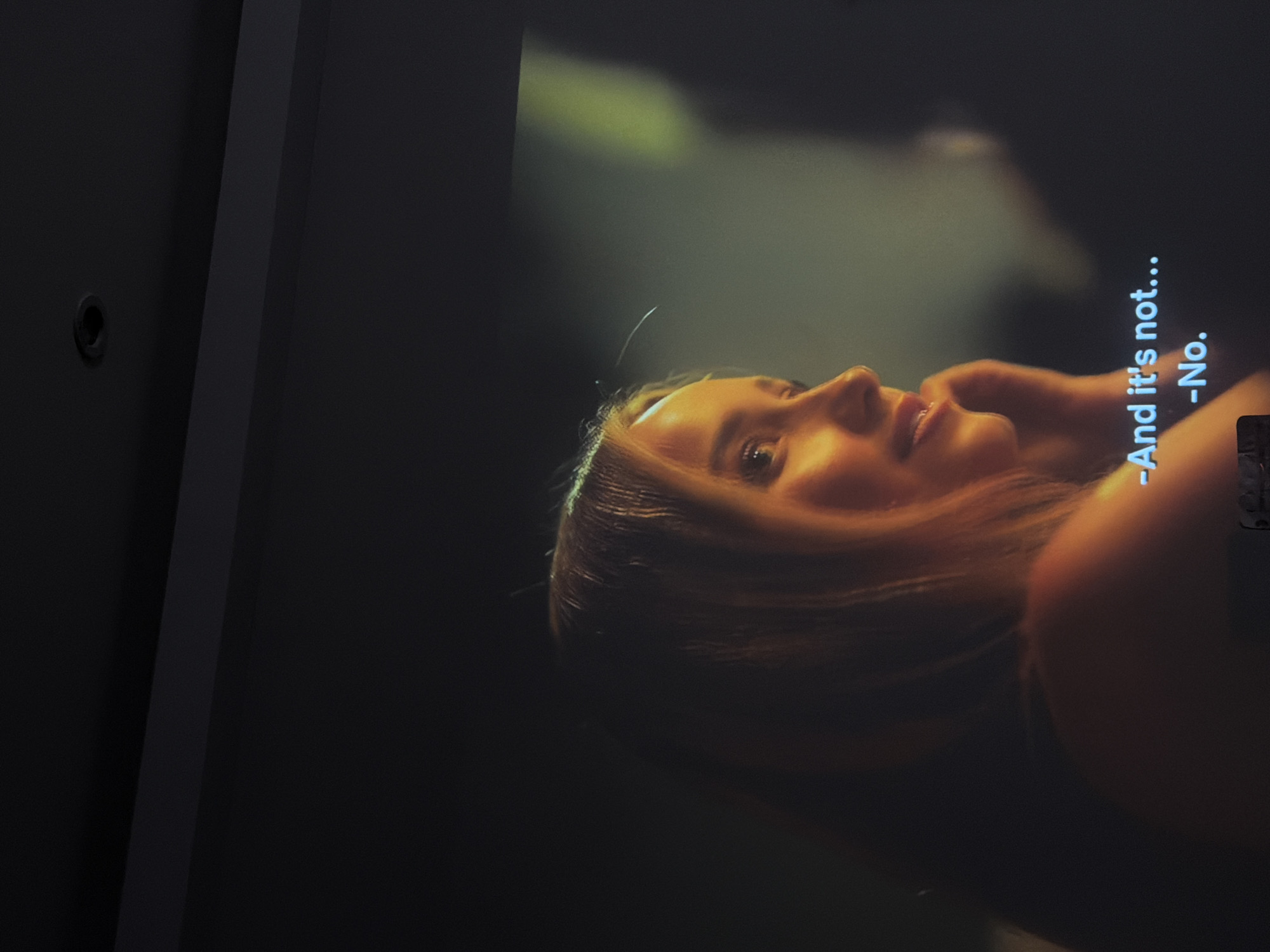 The image is a photograph of a television screen displaying a scene from a film or TV show. The screen shows the profile of a woman with blonde hair, illuminated by warm, golden light. She is looking to her right, with her mouth slightly open as if speaking. There are subtitles visible at the bottom of the screen that read "-And it's not..." and "-No." The background of the screen is dark, with a greenish hue on the right side, suggesting a dimly lit environment. The top portion of the image shows a dark ceiling with a recessed light fixture. There is no indication of the location being in the city of London, United Kingdom.