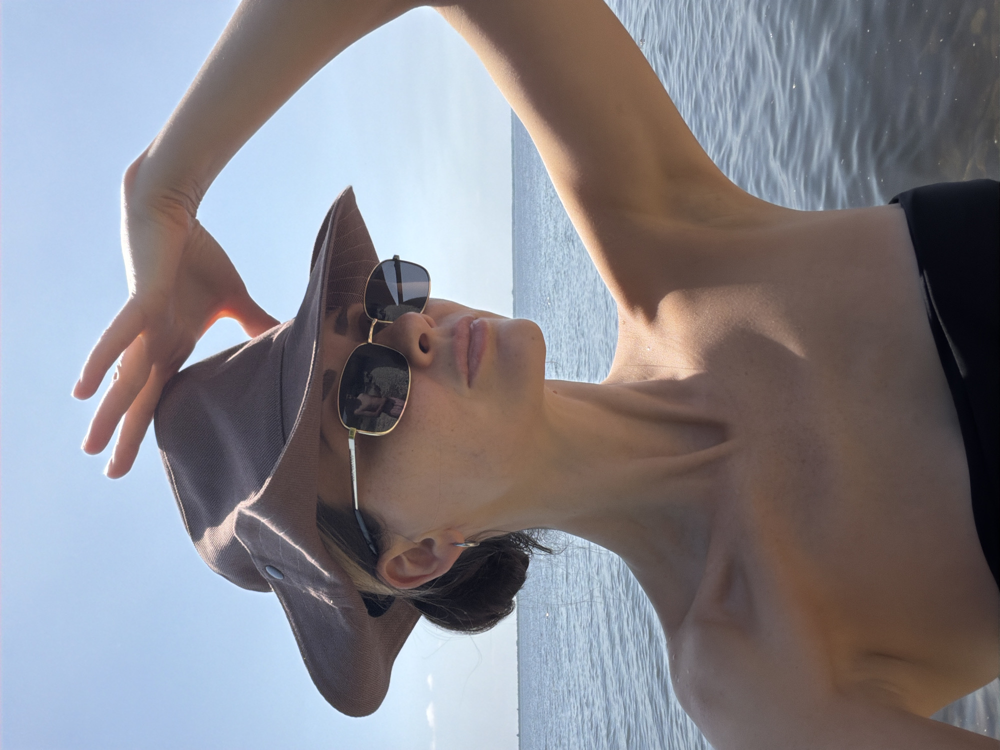 A woman in a brown hat and sunglasses stands by the water, her hand raised to shade her eyes. The water appears calm and the sky is clear blue. The woman is wearing a black top, and her shoulders and neck are visible. The scene suggests a sunny day at a waterfront location. The location in the city of Kamyshin, Russia is not discernible from the image.