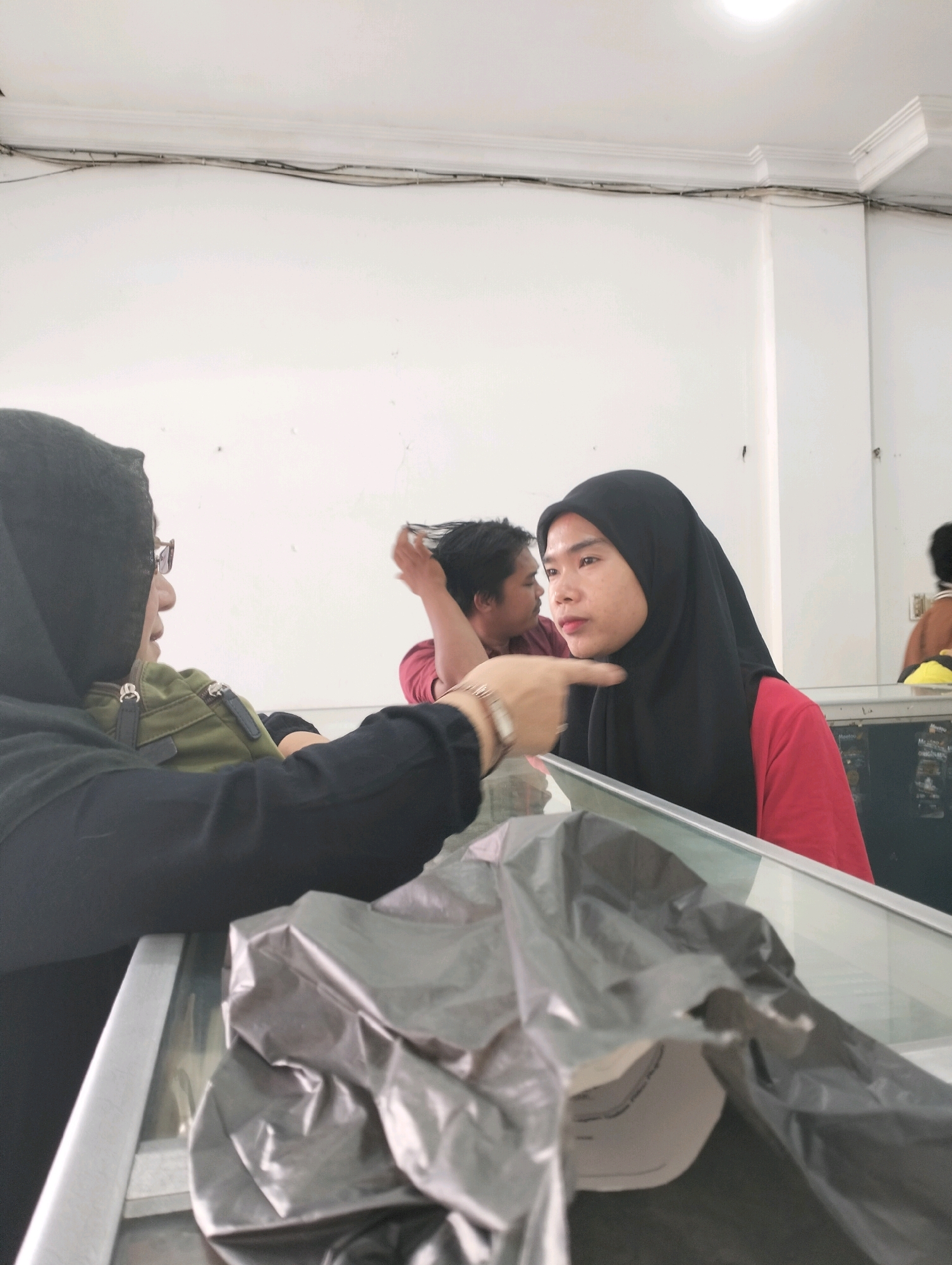 The image shows a group of people inside a building. In the foreground, a person wearing a black hijab and glasses is pointing their finger towards another person. This second person is also wearing a black hijab and a red shirt. Behind them, another person with dark hair is visible, and further back, other individuals are present, some partially obscured. The environment appears to be a retail or service space, with a display counter and possibly goods on shelves in the background. The walls are painted white, and electrical conduits are visible along the ceiling. The lighting suggests an indoor setting. The image is taken from a slightly elevated angle. Without specific visual cues from the image itself, it is impossible to pinpoint the exact location within Bandar Lampung, Indonesia.