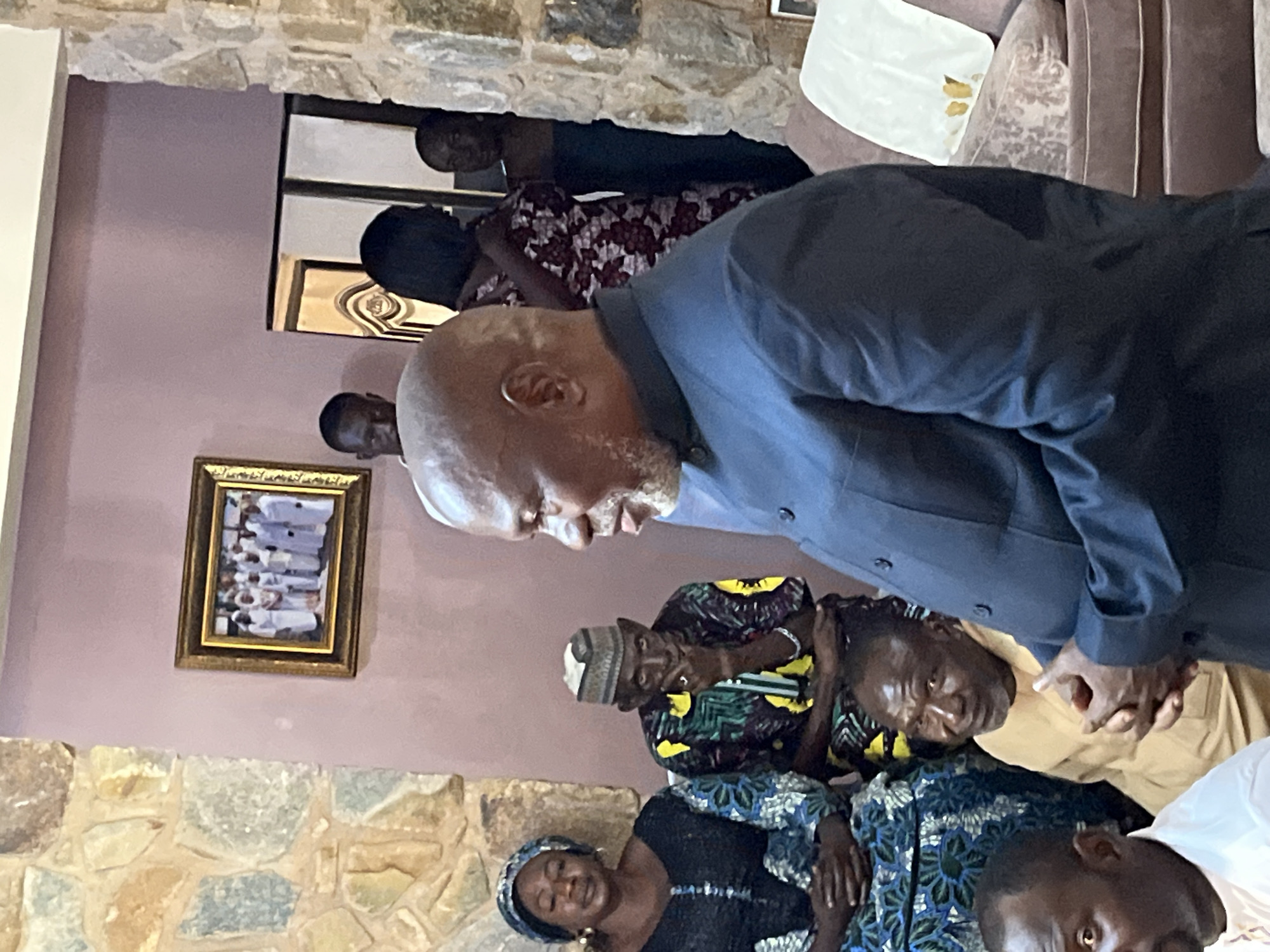 In a room, likely in Bukuru, Nigeria, with stone and mauve walls, several people are gathered. In the foreground, a man with a bald head and grey beard, wearing a dark blue suit jacket, is facing to the right, speaking. To his left, a woman in a blue and black patterned outfit and headscarf is seated, looking down. Beside her, a man in a colorful patterned shirt and a patterned cap is seated, with his hand to his chin. In the background, a framed photograph of people in white robes hangs on the wall. Other individuals are visible in the background, including a woman in a floral dress and two men. The scene appears to be an indoor gathering.
