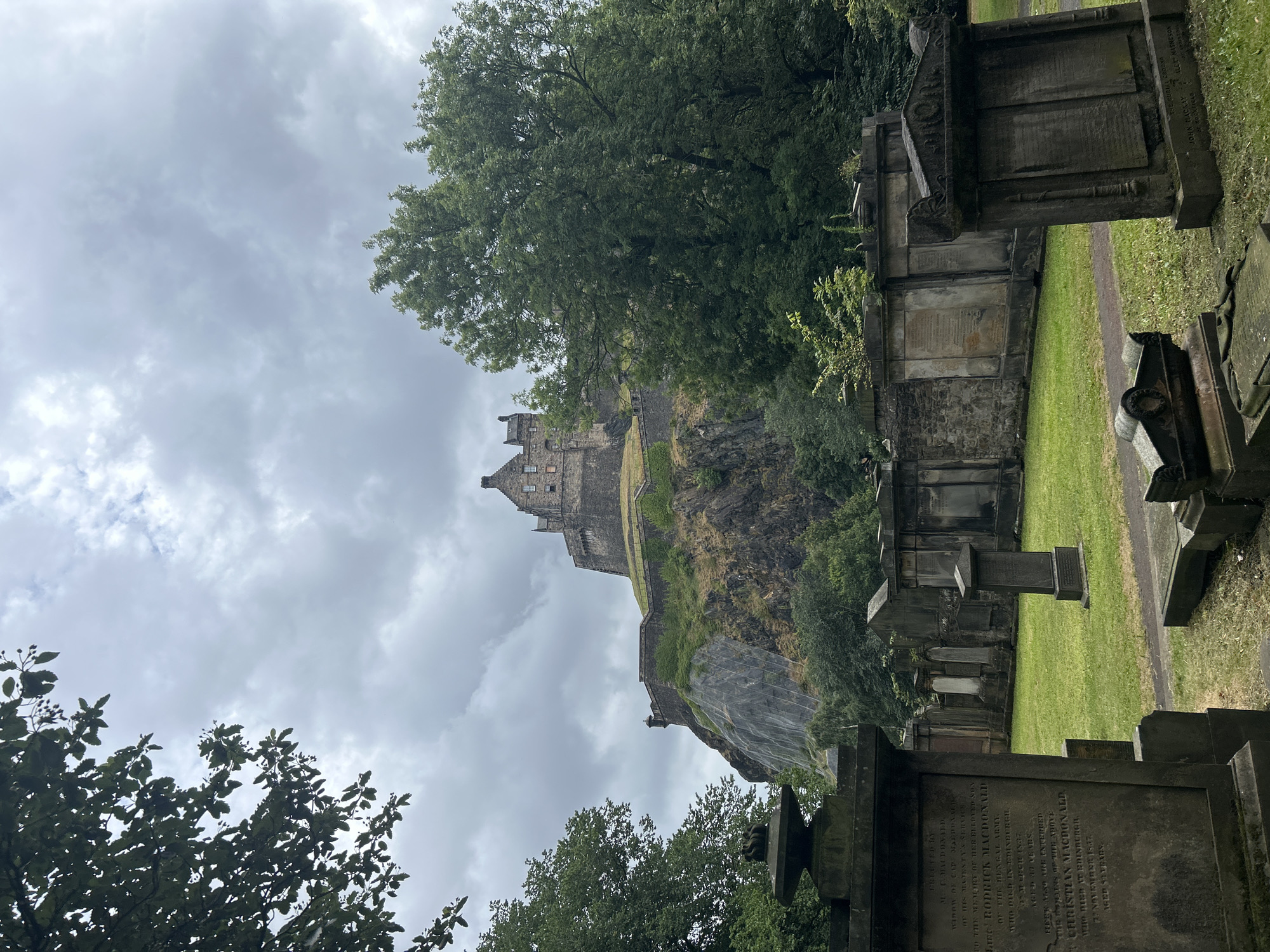 The image depicts a view of Edinburgh Castle, a historic fortress, situated atop Castle Rock in Edinburgh, Scotland, United Kingdom. In the foreground, a graveyard is visible, with several weathered gravestones and mausoleums. Green grass covers the ground between the monuments. Lush trees frame the scene on the left and right, with foliage partially obscuring the sky, which is overcast with grey clouds. The castle itself is a large stone structure with multiple towers and buildings, perched on a steep, rocky incline. A stone wall, covered in greenery, encircles part of the castle's base. The overall impression is one of historical significance and natural beauty under a muted sky.