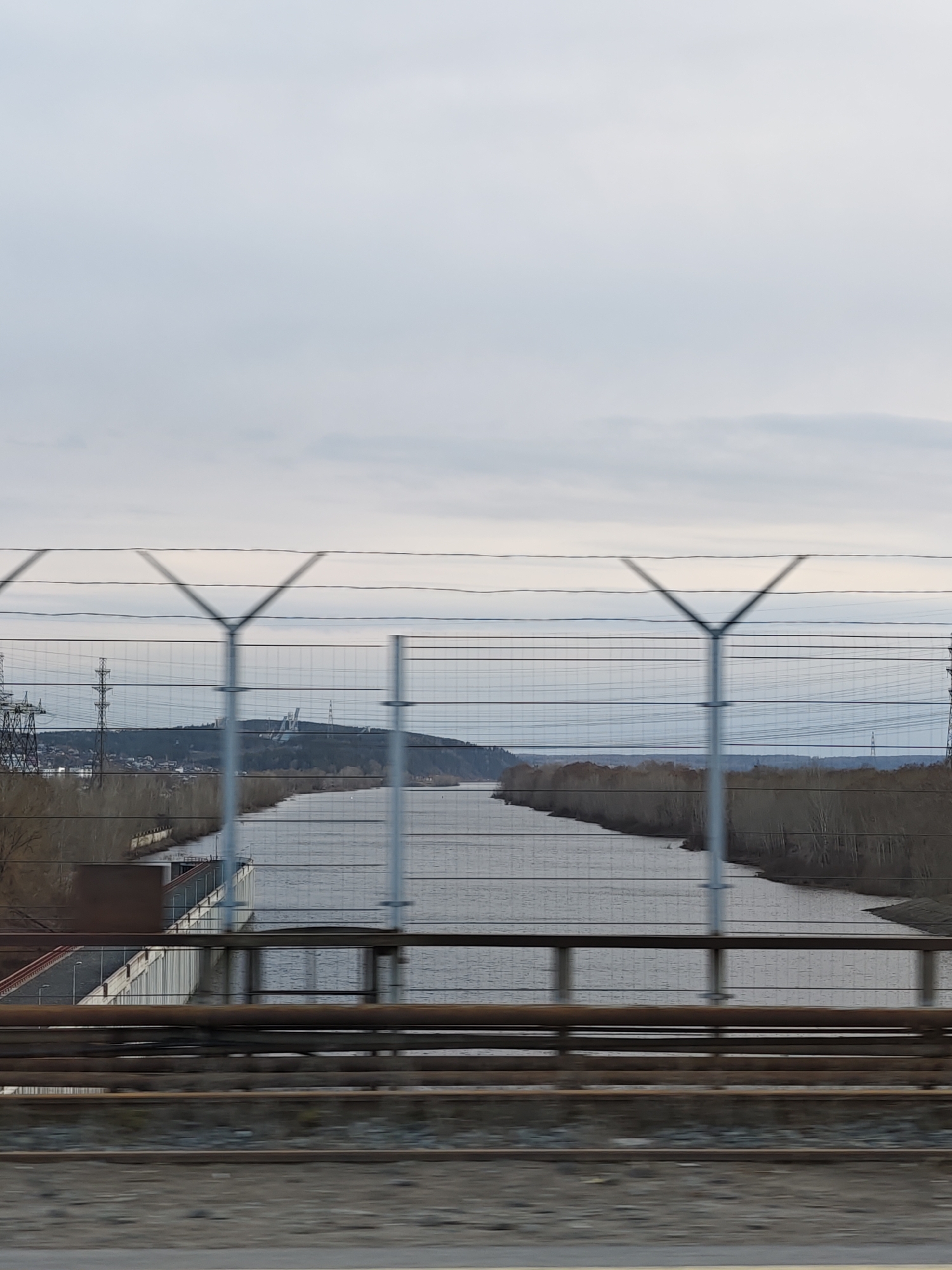 The vertical image presents a wide river or canal flowing through a natural and industrial landscape, viewed from a moving perspective. In the immediate foreground, blurred railway tracks and several horizontal pipelines or structural beams suggest the vantage point is from a train or bridge. A prominent wire mesh fence, featuring distinct Y-shaped support posts, occupies the mid-ground, partially obscuring the scene beyond but allowing a clear view through its grid.
The river itself is a muted gray, reflecting the overcast sky. Its banks are lined with bare trees, indicating a late autumn, winter, or early spring season. On the left bank, closer to the viewer, a concrete retaining wall or industrial structure runs along the water's edge, with some low-lying buildings visible behind it.
In the distance, undulating hills covered with a sparse forest rise above the river. On these hills, various human-made structures can be seen: tall power transmission towers on the left side and a cluster of larger, distinct industrial or architectural buildings atop the central hill. The sky is uniformly overcast, a soft, light gray, suggesting daytime without direct sunlight. There are no people visible, nor any explicit activities taking place. The blur in the foreground suggests motion. No discernible text is visible. The scene depicts a landscape in Chaikovsky, Russia.