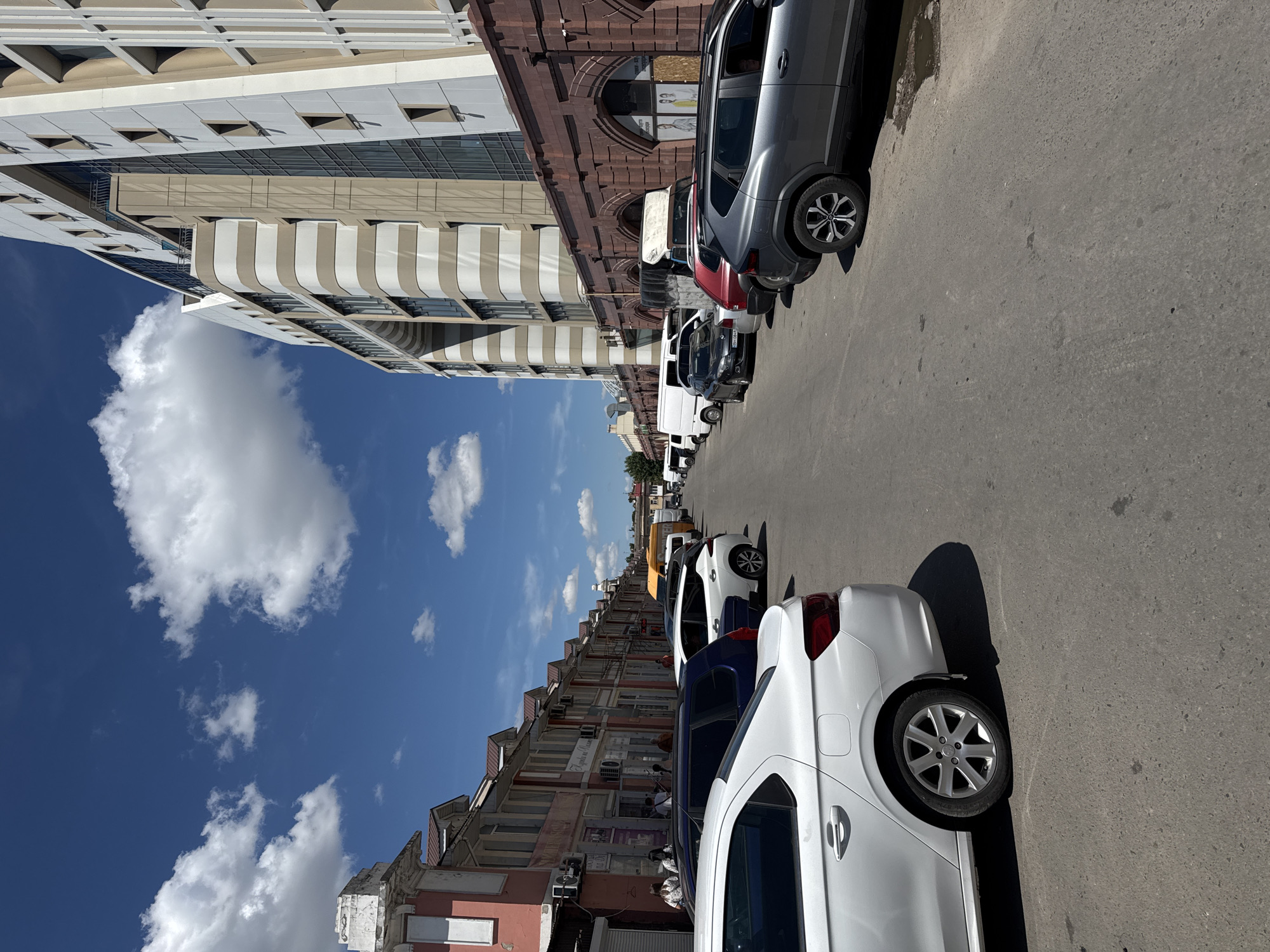 The image shows a street scene in Odesa, Ukraine, on a sunny day with scattered clouds. The street is lined with parked cars on both sides, with a multi-story building on the right and a row of older buildings on the left. The building on the right is modern, with a facade featuring horizontal beige and white bands and large windows. The older buildings on the left have a pinkish hue with pitched roofs. People are visible walking and standing near the buildings on the left. The street appears to be an urban environment with a mix of older and newer architecture.