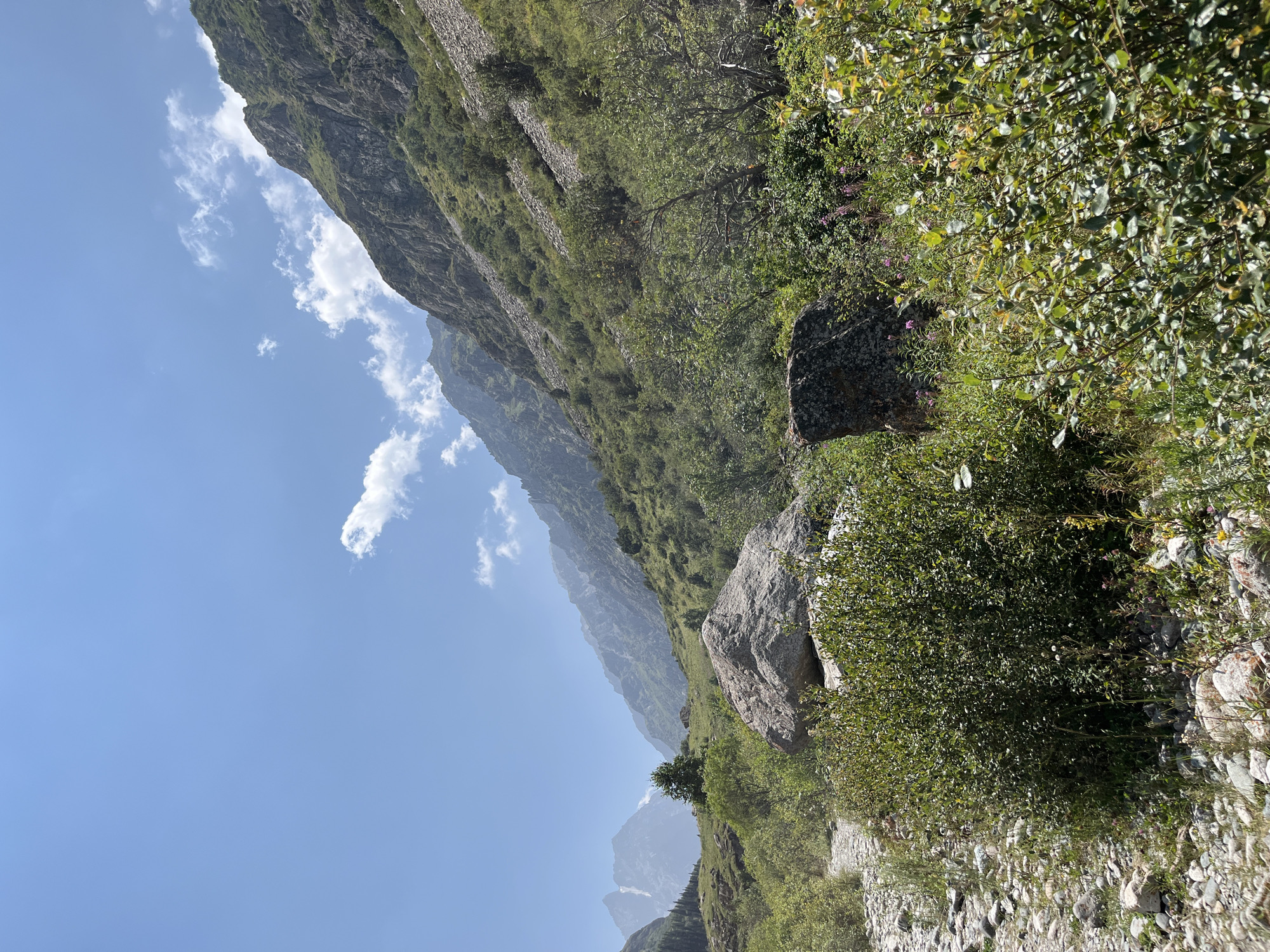 A wide shot captures a mountainous landscape under a clear blue sky with scattered white clouds. In the foreground, a rough, rocky path leads into a verdant valley dotted with green bushes and large rocks. To the right, a steep, rocky slope is covered in sparse vegetation. In the distance, several mountain peaks rise, with the furthest ones appearing hazy and mist-covered. The scene is devoid of any people or man-made structures, suggesting a remote, natural environment. The lighting indicates it is daytime, likely late morning or early afternoon, with the sun casting subtle shadows. The overall impression is one of rugged natural beauty and solitude.