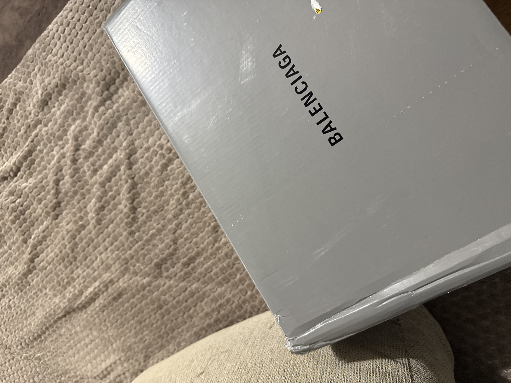 A high-angle, close-up shot shows a light gray Balenciaga box resting on a textured beige surface, likely a cushion or upholstery. The "BALENCIAGA" logo is printed in black, vertically oriented, on the visible top surface of the box. The box shows signs of wear, including scratches and a damaged corner. The background is a tightly woven, textured fabric in a neutral brown tone. The location details are not provided in the image.