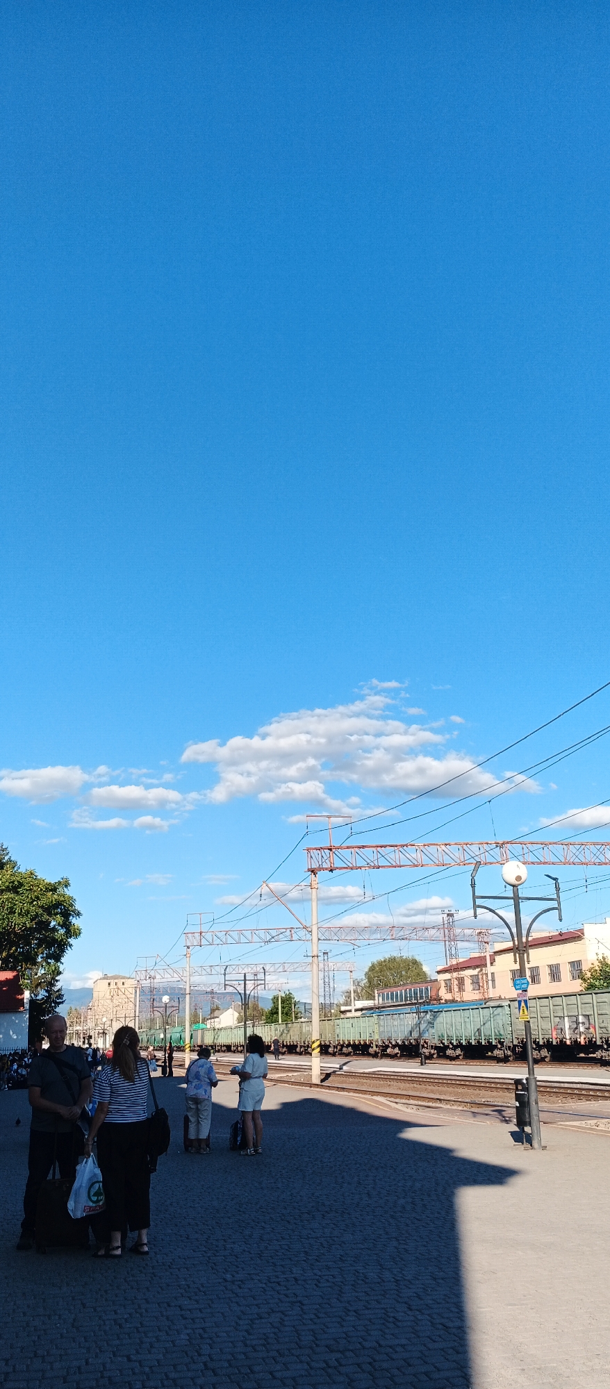 The image depicts a train station platform in Mukachevo, Ukraine. A bright blue sky with scattered white clouds dominates the upper half of the frame. In the lower half, several people are visible, some with luggage, standing on a cobblestone platform. To the right, a train composed of green cargo cars is parked on the tracks. Overhead, electrical wires for the train tracks crisscross the sky. The scene appears to be a sunny day, with shadows cast on the platform by the sun. The architecture visible in the background suggests an urban or semi-urban setting.