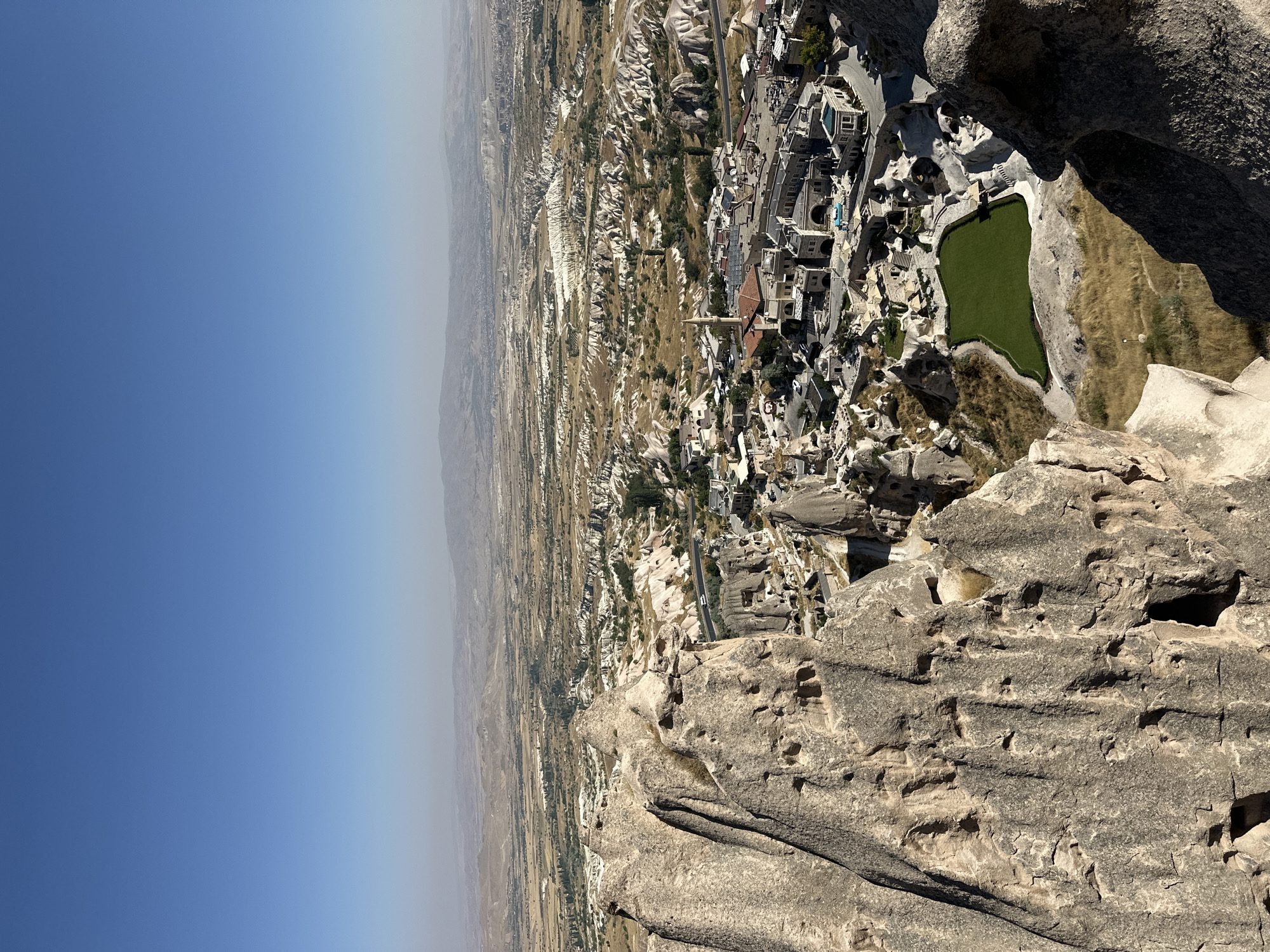 The image captures a panoramic view of Uçhisar Beldesi, Turkey, a town renowned for its unique geological formations and cave dwellings. In the foreground, rugged, light-brown rock formations dominate the left and right sides of the frame, exhibiting a weathered texture with numerous small holes and indentations. These formations obscure parts of the landscape below.
The midground reveals a cluster of buildings, characteristic of the region, with stone facades and a mix of traditional and modern architectural elements. Some structures appear to be carved directly into the rock. A vibrant green, rectangular lawn is centrally located within this built-up area, contrasting with the surrounding earthy tones. A road snakes through the town, with a few vehicles visible. A minaret rises from one of the buildings, indicating a mosque.
The background stretches into a vast, undulating landscape of rolling hills and valleys, dotted with sparse vegetation and more of the distinctive rock formations. The distant horizon is hazy, with mountains faintly visible under a clear, bright blue sky. The overall scene is one of a settlement integrated into a dramatic, natural environment, bathed in natural daylight. There are no visible people or distinct actions taking place.