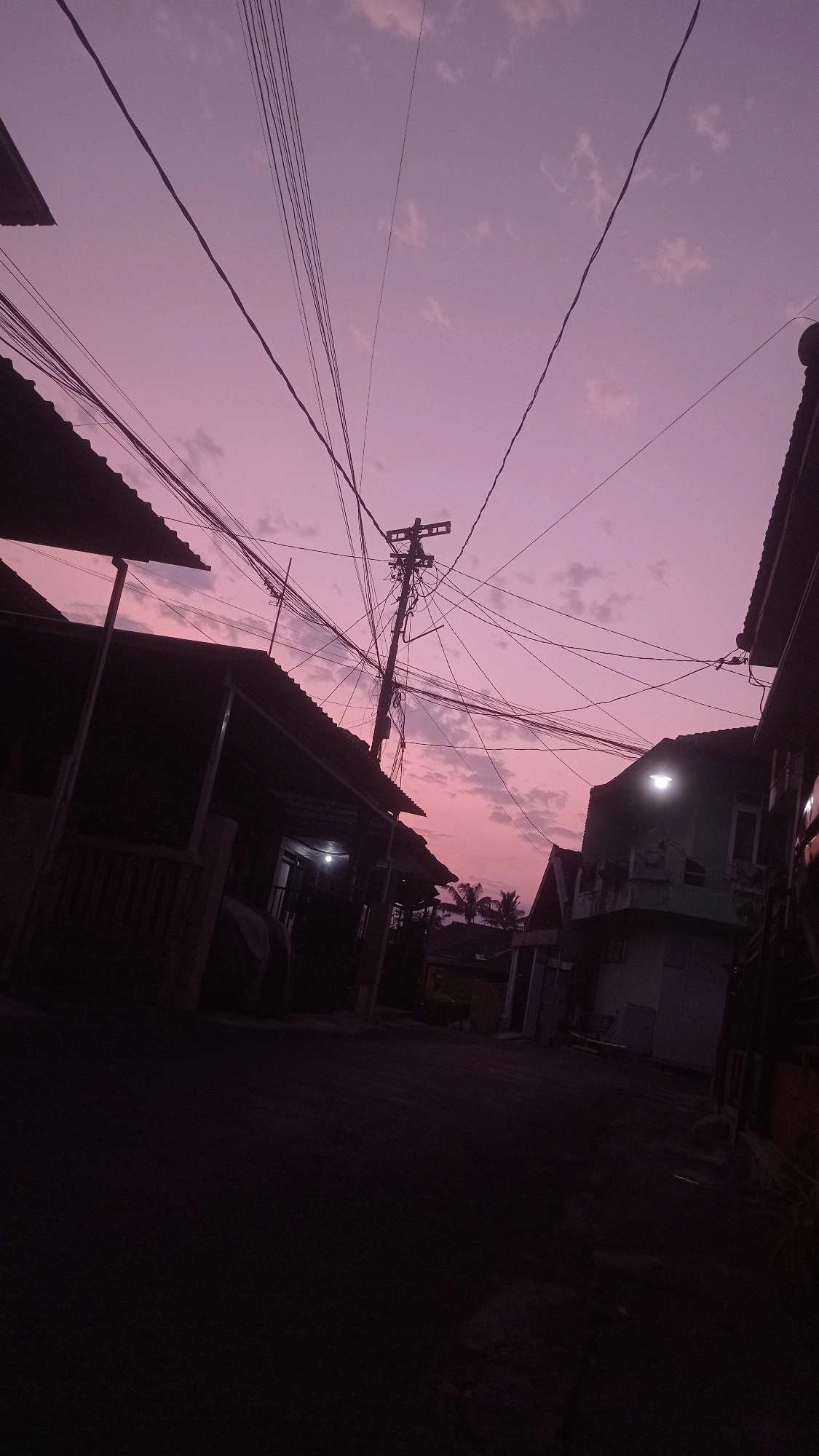 The image depicts a street scene in the city of Mulyoagung, Indonesia, at dusk. The sky is a prominent shade of purple and pink, with scattered clouds, indicating the transition from day to night. A tangle of power lines crisscrosses the sky, suspended from utility poles. In the foreground and midground, dark silhouettes of houses and buildings line the street, their roofs and structures barely visible against the darkening sky. Some buildings have illuminated windows or outdoor lights, casting a faint glow. Palm trees can be seen in the background, adding to the tropical setting. The street itself is unlit and appears to be paved. There are no people visible in the image. The overall mood is quiet and slightly melancholic due to the dim lighting and the stark silhouettes.