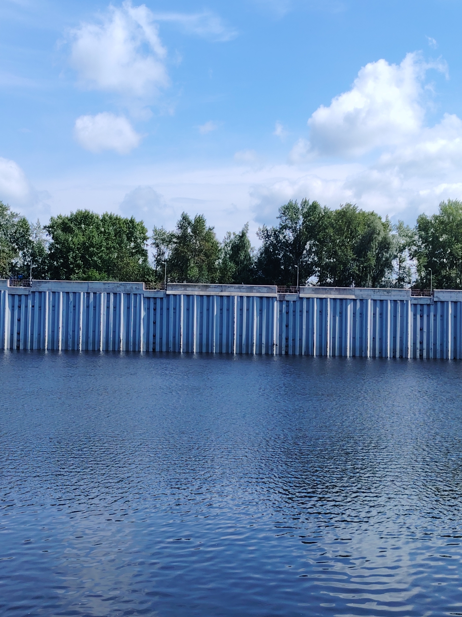 The image captures a waterfront scene in the city of Chaikovsky, Russia, likely along the Kama River. A modern concrete retaining wall with vertical metal sheeting forms a barrier along the river's edge. The wall is topped with a walkway or platform, lined with metal railings. Beyond the wall, a dense line of lush green trees stands against a bright blue sky dotted with fluffy white clouds. The water in the foreground is a deep blue, rippling with small waves, reflecting the sky and surrounding elements. No people are visible in the scene. The overall impression is one of a well-maintained urban waterfront, blending natural elements with industrial infrastructure under a clear, sunny sky.