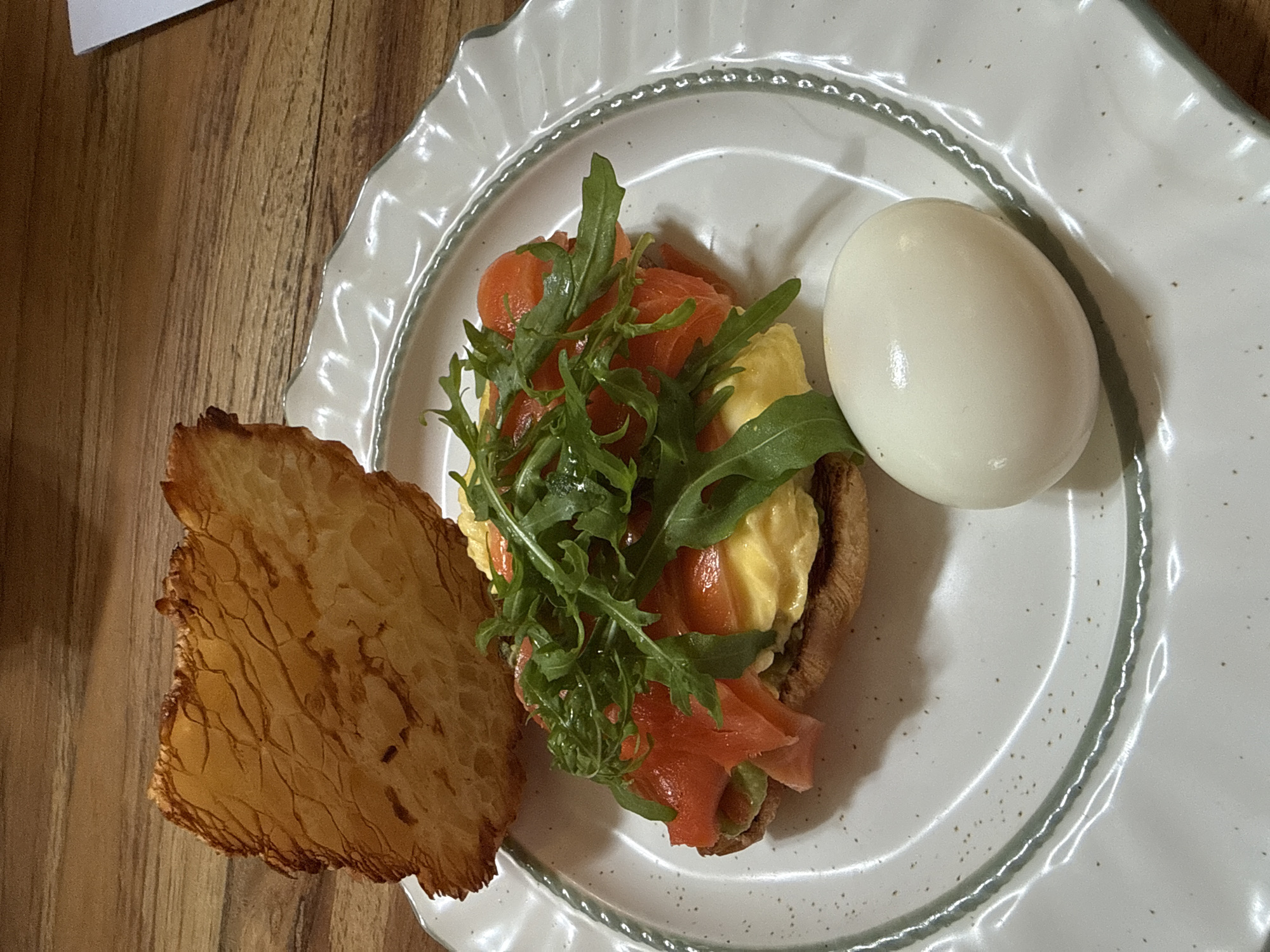 The image depicts a plate of food, likely photographed indoors on a wooden surface. The plate is white with a textured rim, and the food consists of a piece of toasted bread topped with avocado, scrambled eggs, slices of salmon, and arugula. A whole, hard-boiled egg sits beside the toast. There is also a small piece of fried bread or toast. The setting appears to be Ubud District, Indonesia.