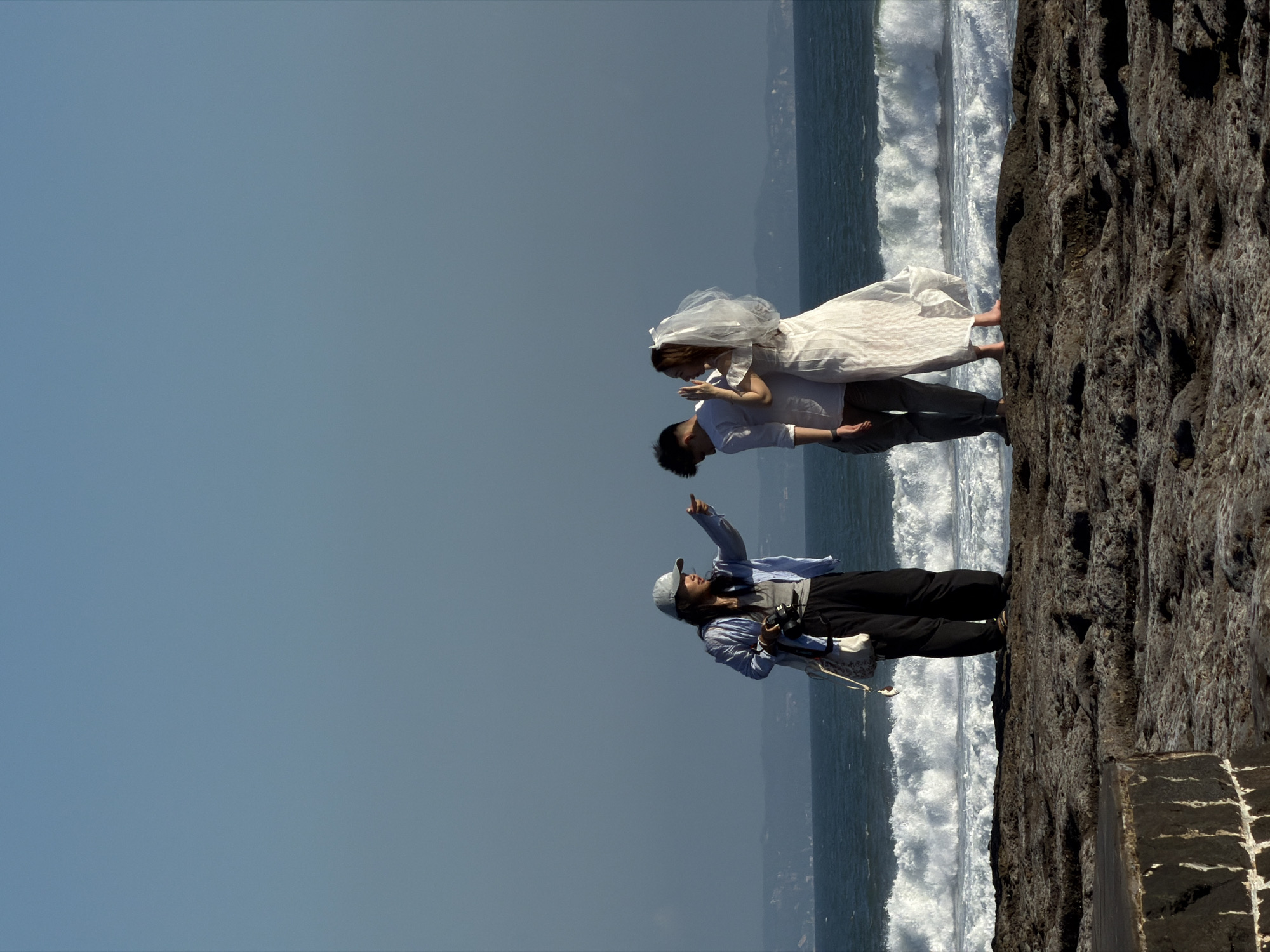 A group of three people are standing on a rocky shoreline with waves crashing in the background. The setting appears to be a beach in Mengwi, Indonesia. On the left is a woman wearing a light blue jacket, grey t-shirt, black pants, and a baseball cap. She is holding a camera in her left hand and pointing to the right with her right hand, seemingly directing the other two individuals. In the center and on the right are a man and a woman, presumably a couple. The man is wearing a white long-sleeved shirt and grey pants. The woman is wearing a white wedding dress and a veil, her back is partially turned to the camera as she looks towards the man. The ocean is a deep blue with white foamy waves. The sky is clear and a light blue. The ground they are standing on is composed of dark, rough rocks.