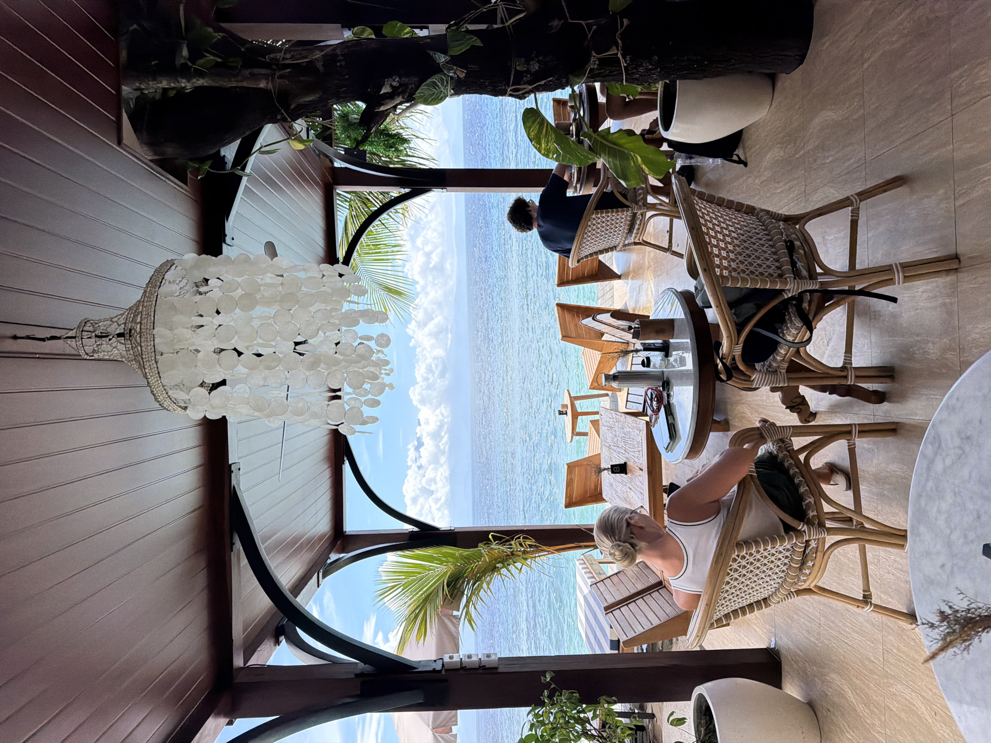 A sideways view depicts an outdoor dining scene in Nusa Penida, Indonesia. Two people sit at tables overlooking a sea. A woman with blonde hair sits in a wicker chair at the right. A person sits at another table further away. The tables are round and small. The view to the sea is open, revealing clear water and a light blue sky with clouds. Lush green plants and palm trees frame the scene. Overhead, a unique shell chandelier hangs from the wooden structure. White planters holding plants stand in the foreground and background.