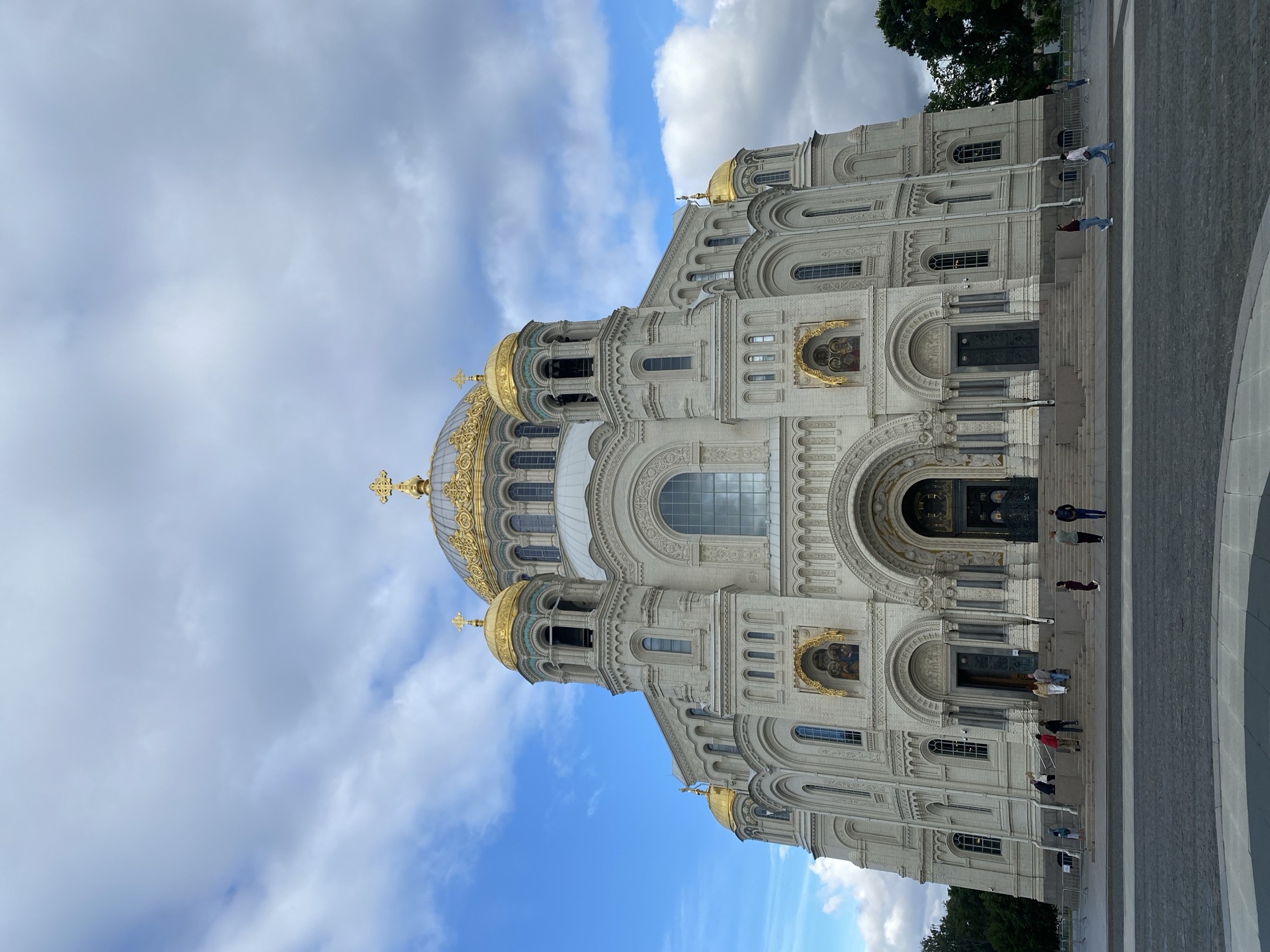 The image shows a wide-angle, low-angle exterior view of the Kronstadt Naval Cathedral in Kronstadt, Russia. The cathedral is a large, white, multi-domed structure with golden cupolas topped with crosses. The facade features multiple arched entrances and windows, as well as decorative gold elements. There are several people walking on the steps and on the plaza in front of the cathedral. The sky is cloudy with patches of blue sky visible. To the right of the cathedral, a dense cluster of green trees is visible. The ground in front of the cathedral is paved with cobblestones.