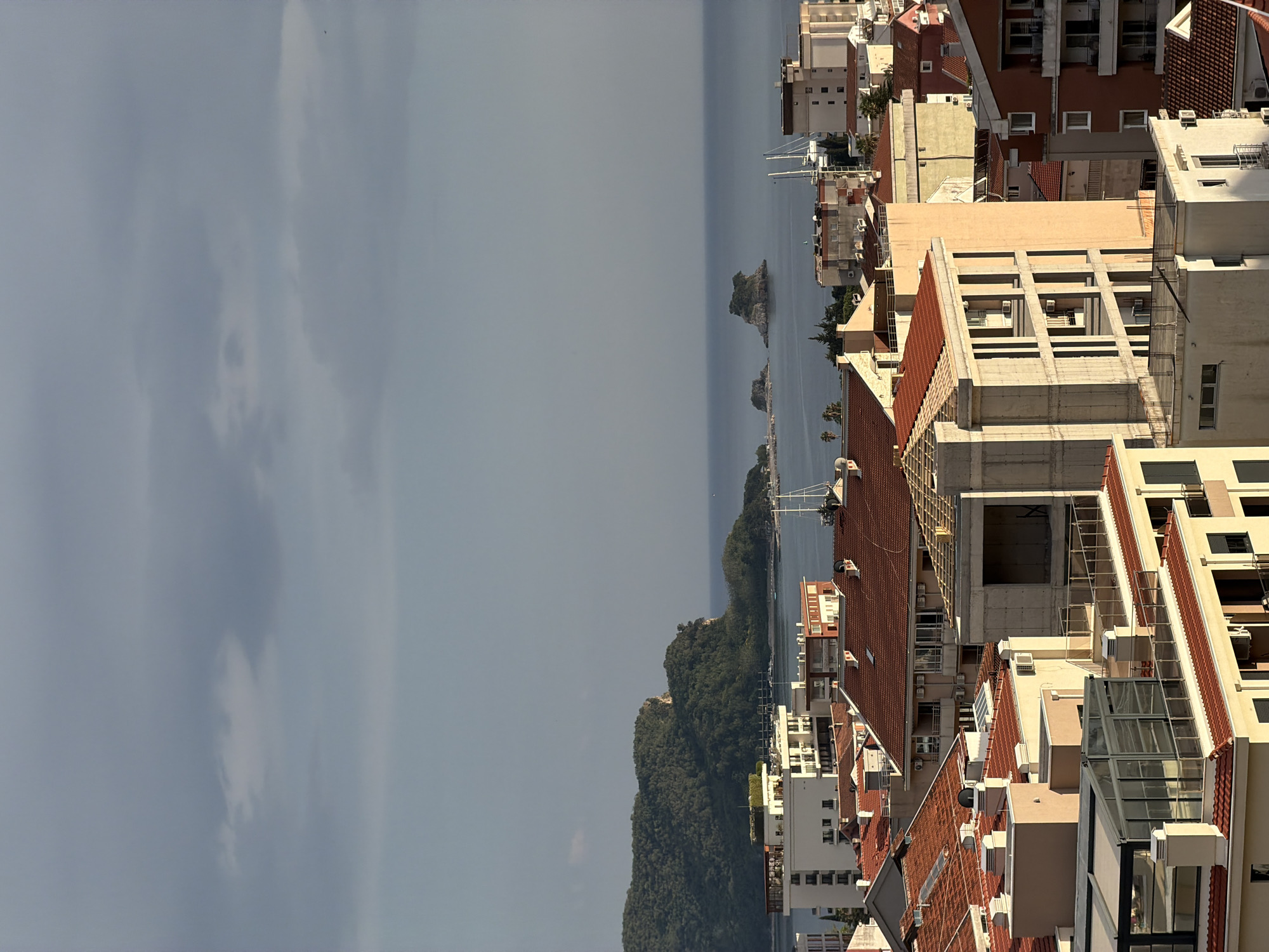 The image shows a coastal view of Budva, Montenegro. The right side is dominated by buildings with red-tiled roofs and concrete structures. The buildings appear to be multi-story apartments or hotels. The center of the image shows a body of water with a small island. On the left is sky filled with clouds. The buildings are positioned on a slight slope, and the landscape appears to transition from dense vegetation to urban development.