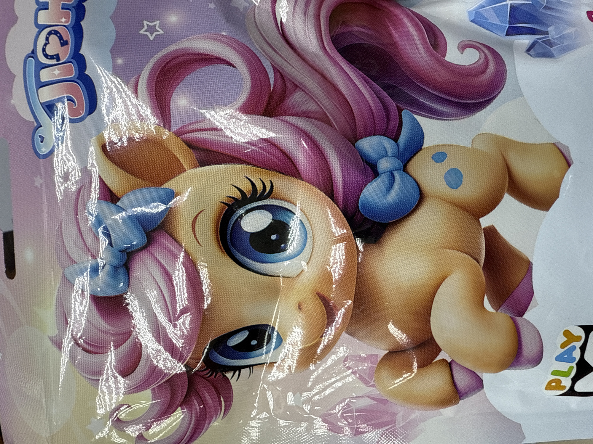 This image shows a close-up of a toy featuring a cartoon pony or horse with a light tan body and pink and white streaked hair styled in swirls. The pony has large, bright blue eyes with black eyelashes. Blue ribbons are tied in its hair. Adjacent to the pony is a second, similar-looking character, also with a tan body and blue markings. The pony is printed on a surface, possibly packaging, which is partly covered by a crinkled transparent material. The background is a pastel lavender with various decorative elements. The word "PLAY" is visible in the lower right corner. There is no indication of this image being in Votkinsk, Russia.