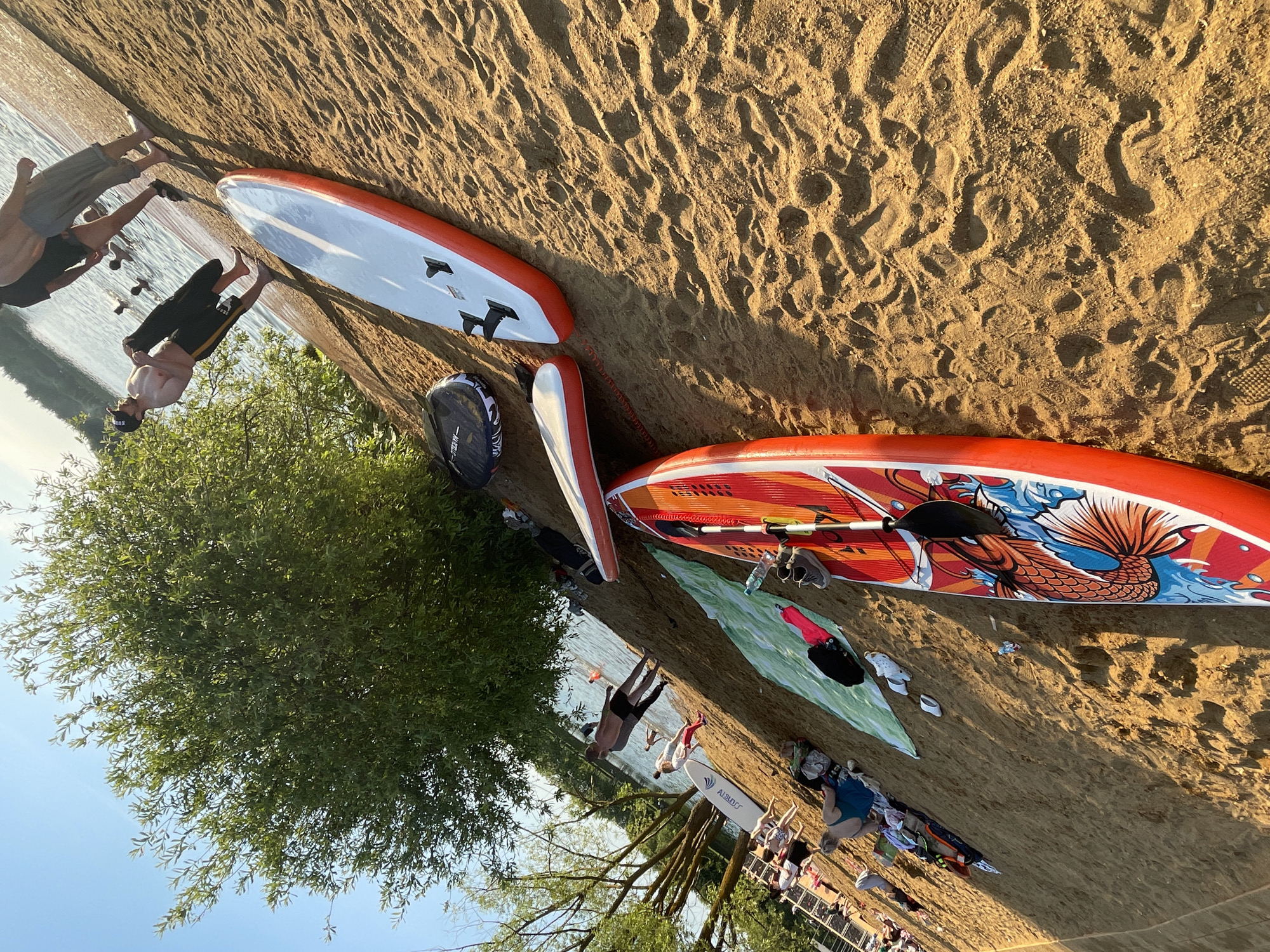This image depicts a sandy beach scene in Moscow, Russia, illuminated by strong sunlight from the upper right, casting long shadows towards the left.
In the foreground, a vibrant red and orange paddleboard (SUP) adorned with a large fish or dragon graphic and blue wave patterns rests horizontally on the sand, with a black paddle lying across it. To its left, a plain white and orange paddleboard is partially visible. Further back, a third white paddleboard with "AISBERG" branding is also on the sand. A black and white circular object is positioned near the plain white and orange SUP. The light brown sand is imprinted with numerous footprints and shoe marks. A green towel is spread out, holding a dark bag, a pair of white sandals, and a plastic water bottle. A pair of grey sneakers lies near the red and orange paddleboard.
In the mid-ground, near the water's edge, a shirtless man wearing dark shorts and a dark baseball cap stands, holding a dark, possibly folded garment. Further into the shallow water, at least three other individuals are partially submerged, appearing to be wading or swimming. On the sand to the left, another shirtless man in dark shorts stands near the water.
The background shows a calm body of water, likely a lake or large pond. Along the left side of the beach, a large, leafy green tree with dense foliage provides shade. Further down the beach, multiple people are visible, some lounging on beach chairs, others on towels. White structures, possibly part of a beach facility, are visible on the far left. The sky above is clear and blue.