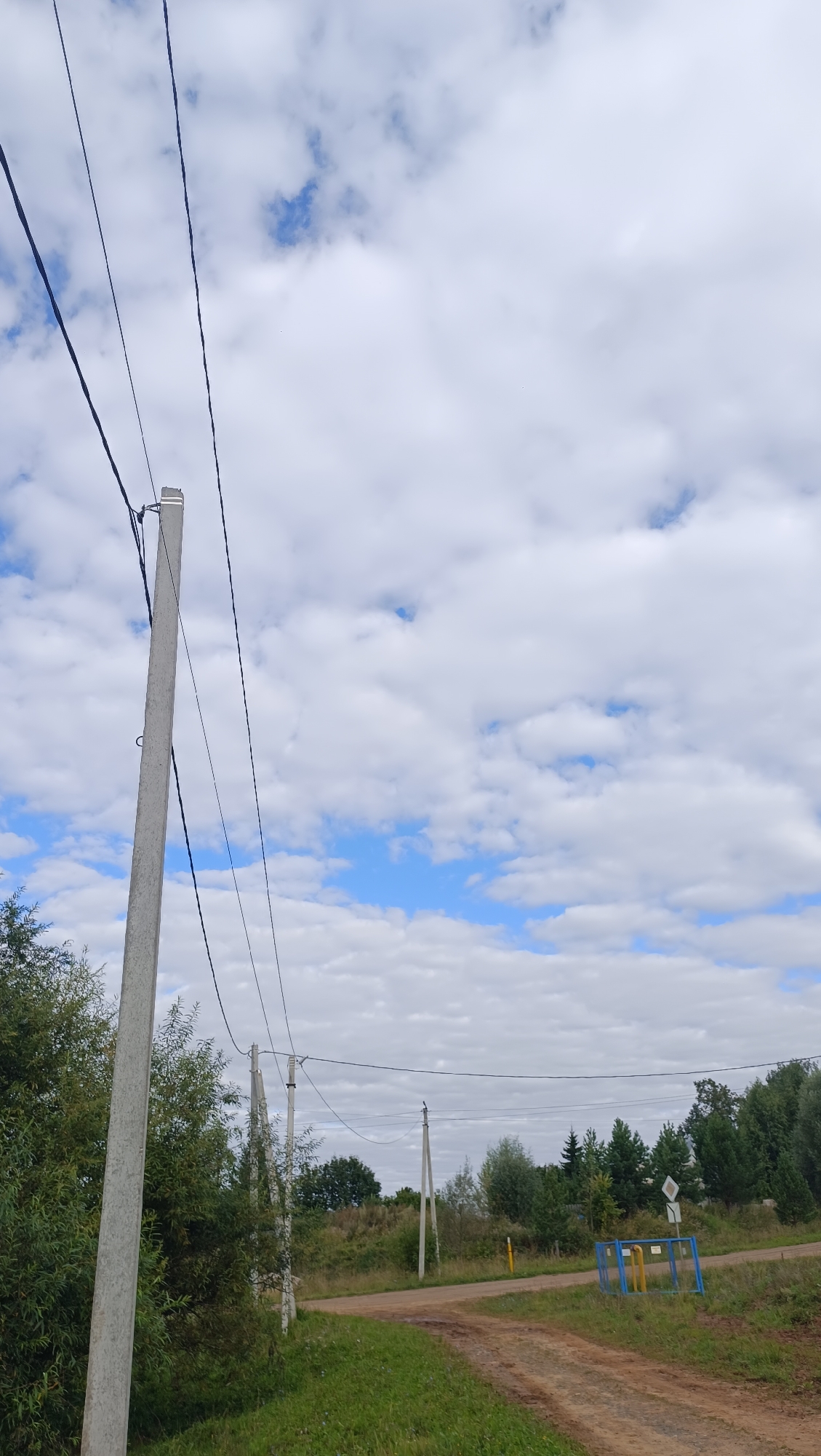 A rural dirt road curves through a grassy landscape under a cloudy blue sky. Several power poles with cables run along the side of the road, with trees and vegetation flanking the path. In the distance, there is a yellow and blue metal structure with signage. The scene appears to be located outside of Karsha, Russia.