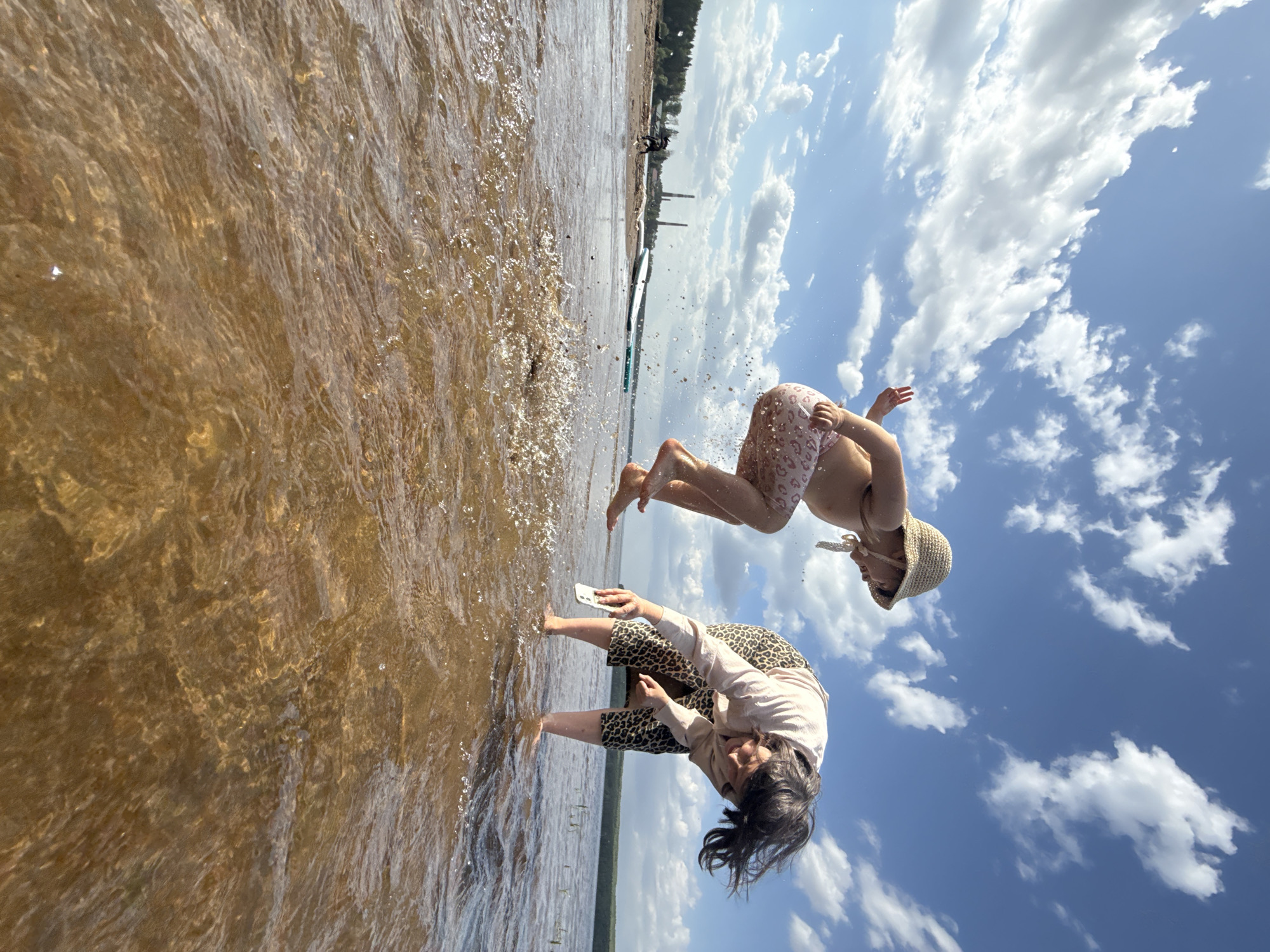 The image shows two people in the water at the beach in Chaikovsky, Russia. One adult female is standing in the water with a phone in her hand. A young girl is jumping out of the water and flipping in the air, wearing a hat. The water is murky. The sky is visible in the background. The beach appears to be along a river or lake. Trees are visible in the distance. A few structures are also seen on the shoreline. The photo is taken at a slight angle, rotated counter-clockwise.
