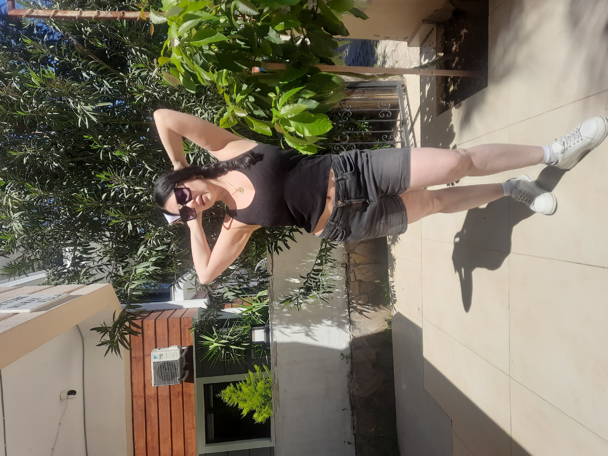 A full-body, portrait-style photograph captures a woman posing outdoors on a sunny day in what appears to be a garden or patio setting, consistent with Kemer, Turkey. The woman stands upright, with her arms raised and hands clasped behind her head in a relaxed pose. She has long dark hair styled in a braid and wears dark sunglasses, a plain black tank top, and dark grey denim shorts. Her footwear consists of white sneakers with white socks. A small gold-colored pendant necklace is visible around her neck.
The background is dominated by lush, dense green foliage from various trees and bushes, suggesting a vibrant natural environment. To the left, part of a building is visible, featuring a light-colored stucco-like wall contrasting with a section of vertical brown wooden siding. A window, an outdoor light fixture, and a wall-mounted air conditioning unit are integrated into the building's facade. A small white dome-shaped security camera is also attached to the building. To the right of the woman, a black metal railing with decorative scrollwork is visible. The ground is a light-colored, tiled surface, where strong, well-defined shadows, including the woman's, indicate bright, overhead sunlight, suggesting mid-day. No legible text is visible in the image.