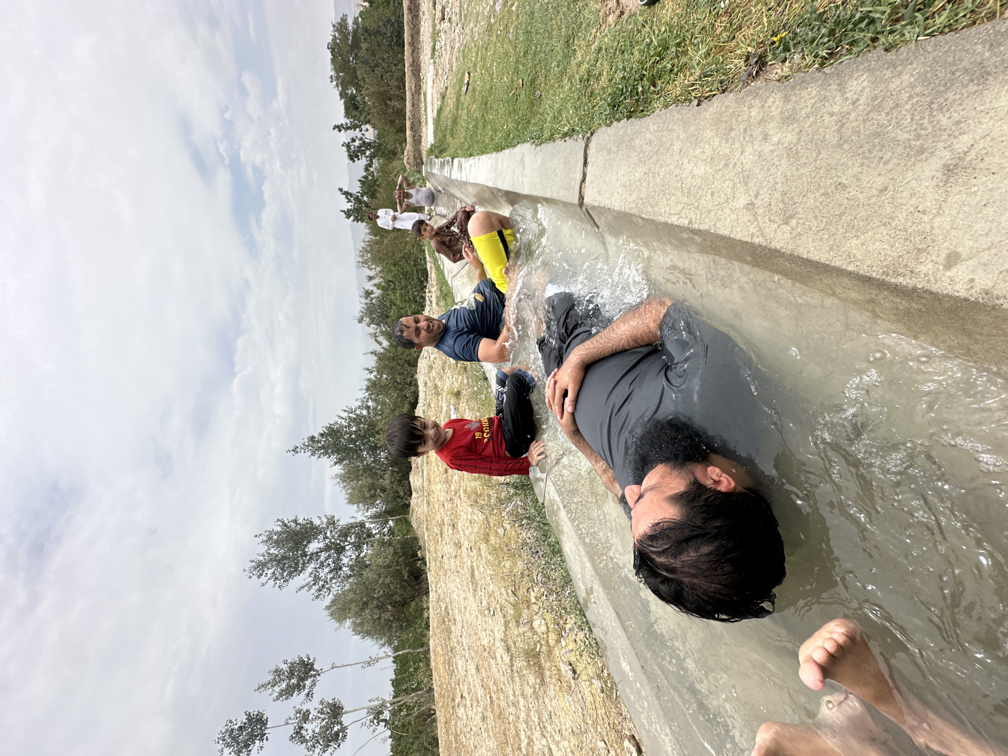 In Kan Mehtarzai, Pakistan, a group of people are enjoying a water channel. The foreground features a man lying on his back in a concrete water channel filled with murky water, his head submerged. His legs and feet are visible above the water. To his left, another man is also partially submerged in the channel, wearing a dark t-shirt and yellow shorts, with a broad smile on his face. A young boy in a red long-sleeved shirt and black pants sits on the edge of the channel, smiling. Further back, a man in a brown outfit sits in the channel, and behind him, two men stand in the distance, one in white traditional attire. The environment consists of a dry, sandy ground with sparse vegetation and trees, and a cloudy sky. The scene appears to be an informal recreational activity, likely during warm weather, utilizing the water channel for cooling off.