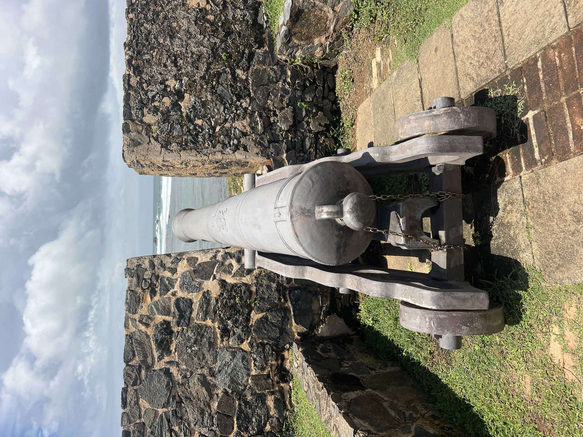 The image shows a historical cannon positioned between two stone walls, overlooking the ocean. The cannon is made of dark metal and rests on a wooden carriage with two large wheels. It is secured by a chain. The stone walls are rough and appear ancient, with grass growing in between the stones and along the top edges. The ground in front of the cannon is paved with stone slabs and some brickwork. Beyond the walls, the ocean is visible with waves breaking on the shore. The sky is partly cloudy with a mix of white and grey clouds. This scene is likely located at the Galle Fort in Galle, Sri Lanka, a UNESCO World Heritage site known for its colonial-era fortifications. There are no people visible in the image. The environment suggests a historical landmark with a view of the sea.