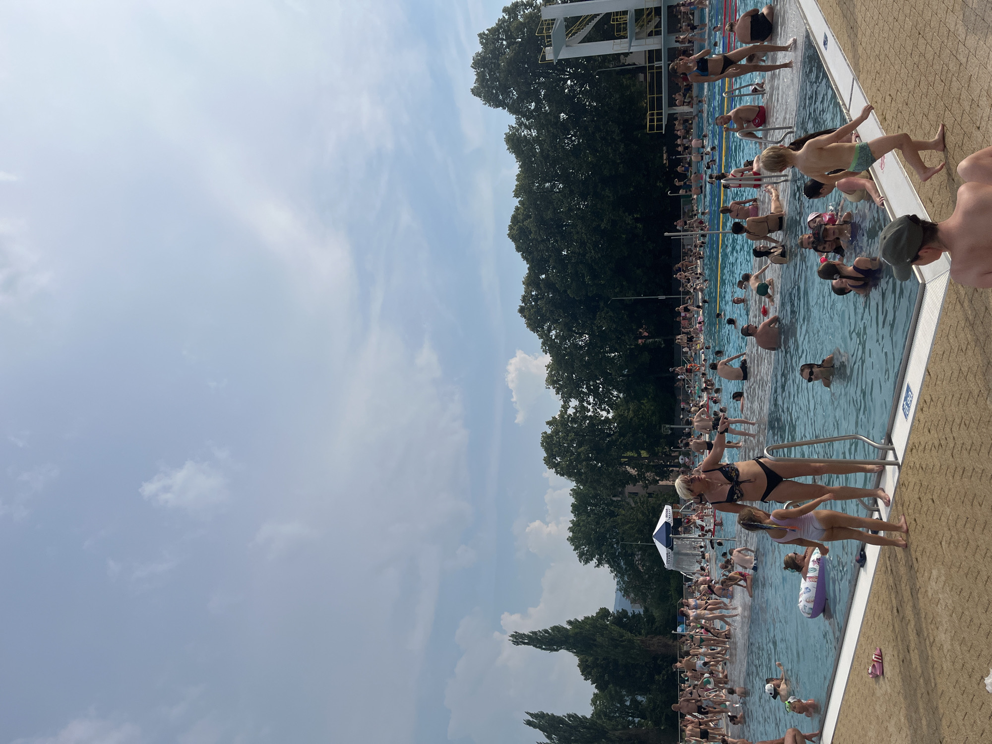 The image depicts a crowded outdoor swimming pool on a sunny day. Numerous people of various ages are in and around the pool, engaged in activities such as swimming, wading, and sunbathing. The pool area appears to be a public facility, with a diving platform visible in the background and lounge chairs lining the deck.
The scene is bustling with activity, suggesting a recreational outing or a popular summer day at the pool. The weather appears to be warm and pleasant, with a clear blue sky and scattered clouds. The time of day is likely midday or early afternoon, given the bright sunlight.
Notable details include a young boy in swim trunks standing at the edge of the pool, a woman in a black bikini posing near a ladder, and children playing with a float. The pool appears to be well-maintained, with clear blue water and clean concrete decking. There are trees in the background, providing some shade and enhancing the outdoor atmosphere.
A small blue and white umbrella is set up near the pool, and a few pairs of flip-flops are visible on the deck. There is a marker on the side of the pool with the text "0.90". The overall mood of the image is one of joy and relaxation, with many people enjoying the summer weather and the amenities of the pool.