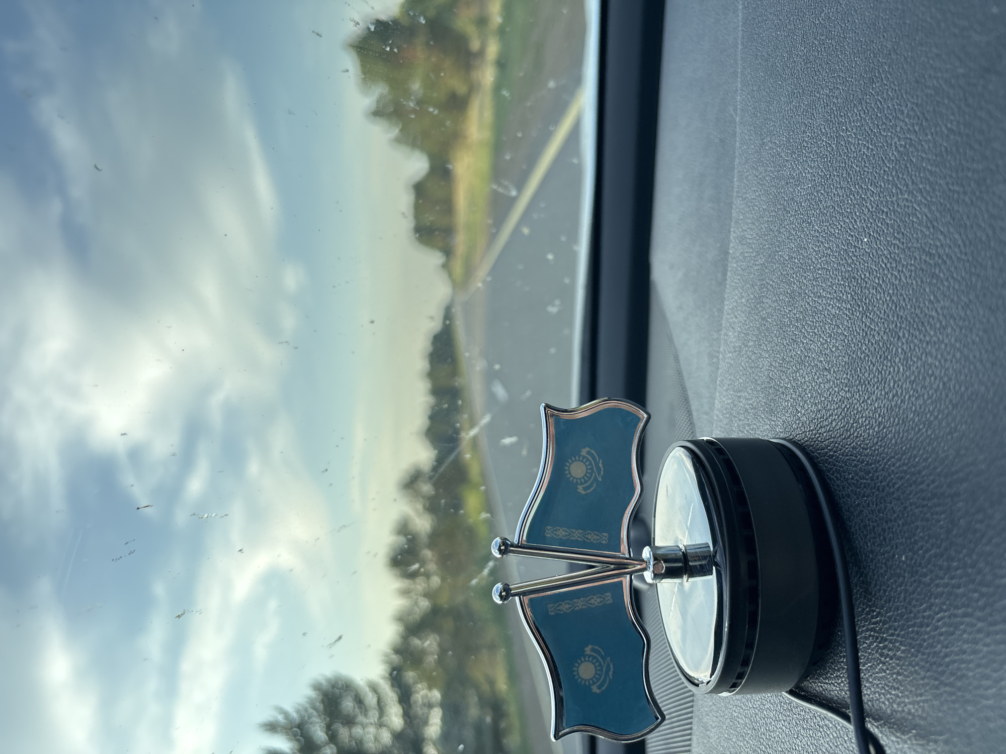 A car dashboard is visible in the foreground, with a dual-flag ornament featuring the flag of Kazakhstan prominently displayed. The flags are attached to a silver stand, which is mounted on a black base with a thin black wire extending from it. The dashboard itself has a textured, dark gray surface.

In the background, a road stretches into the distance, bordered by trees and vegetation. The road appears to be a highway with a yellow line marking the lanes. The sky above is partly cloudy with a soft, hazy light, suggesting it might be early morning or late afternoon. The scene is viewed from inside the car, looking through the windshield, which has some dust or debris on it.

The image is set in Глухов ауылдық округі, Kazakhstan.