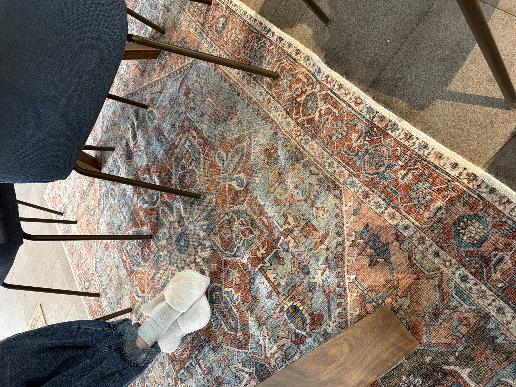 A high-angle shot depicts a portion of a room. A dark blue chair sits on the left side of the frame, with its legs extending onto an intricate area rug. The rug features a blend of blue, brown, and rust-colored patterns. A pair of white socks worn with dark blue jeans are visible in the lower-left corner of the image. The room appears to have a hard floor, possibly tile or concrete. The image does not give enough information to determine if the photo was taken in Chkalov, Armenia.