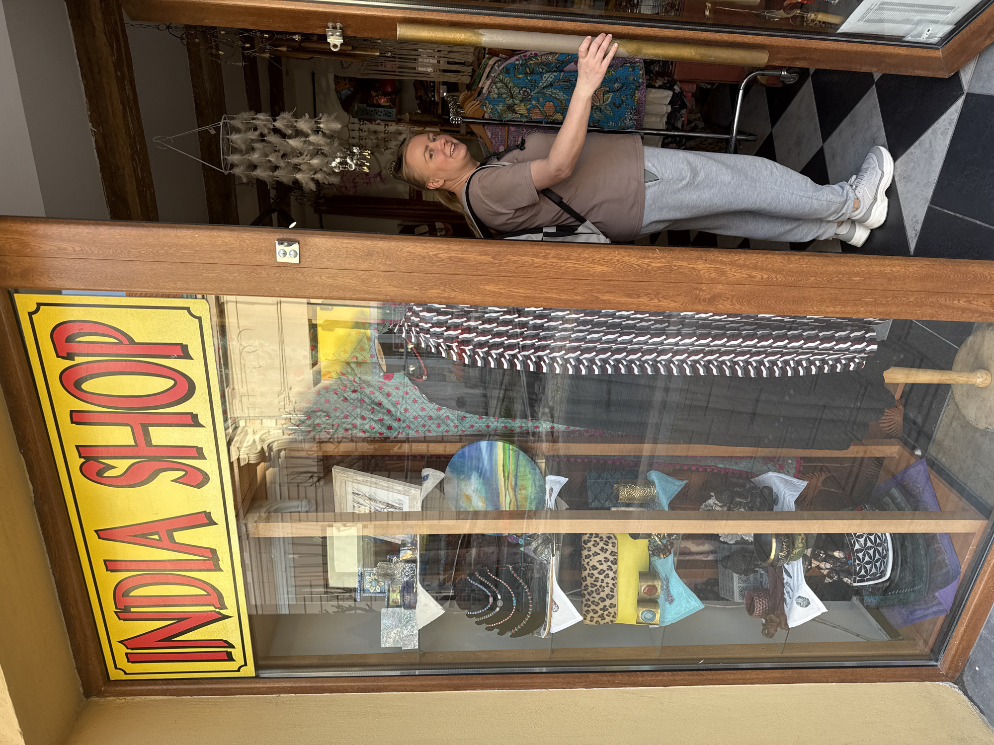 A woman in a brown T-shirt, gray sweatpants, and white sneakers stands in the doorway of an "INDIA SHOP" store in Unknown, Unknown. She has a backpack on her shoulders. The store window displays clothing, jewelry, and decorative items on shelves. A sign on the door reads "INDIA SHOP" in red letters on a yellow background. The flooring inside the shop is black and white checkered. A feather chandelier hangs from the ceiling.