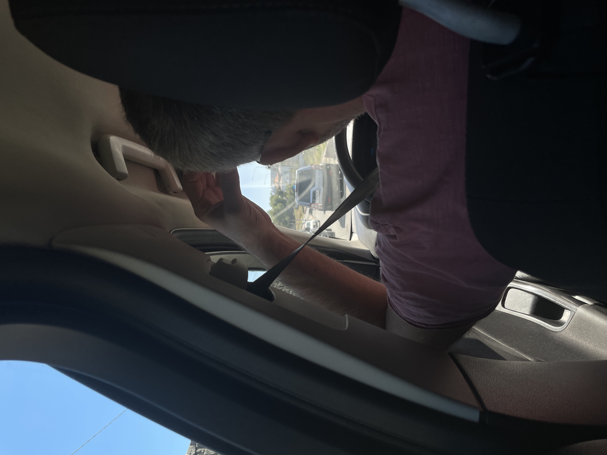 An interior view of a vehicle in Rostov-on-Don, Russia, shows a man driving. He is wearing a pink shirt and has gray hair. The man is adjusting something on his head with his right hand, while his left hand is on the steering wheel. A seatbelt is visible across his chest. Through the car window, other vehicles can be seen on a road, suggesting traffic. The sky is clear and blue, indicating daytime. The interior of the car includes a door handle and part of the dashboard.