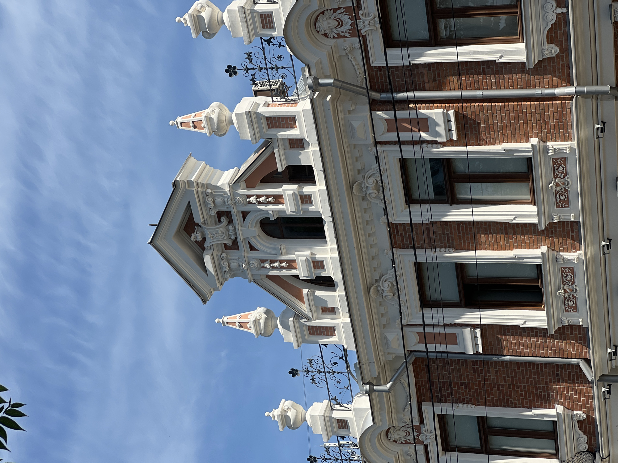 The image shows the upper facade of a historical building with ornate architectural details in Krasnodar, Russia. The building features a brick lower section with several windows, each framed by decorative white stucco work. Above the windows, the facade transitions to more elaborate white plaster ornamentation, including volutes, festoons, and a central pediment with a small attic window. The roofline is adorned with decorative urns and metal railings. The sky above is blue with thin, wispy clouds, and a few green leaves from a tree are visible in the top left corner. The overall impression is one of an old, well-maintained urban structure.