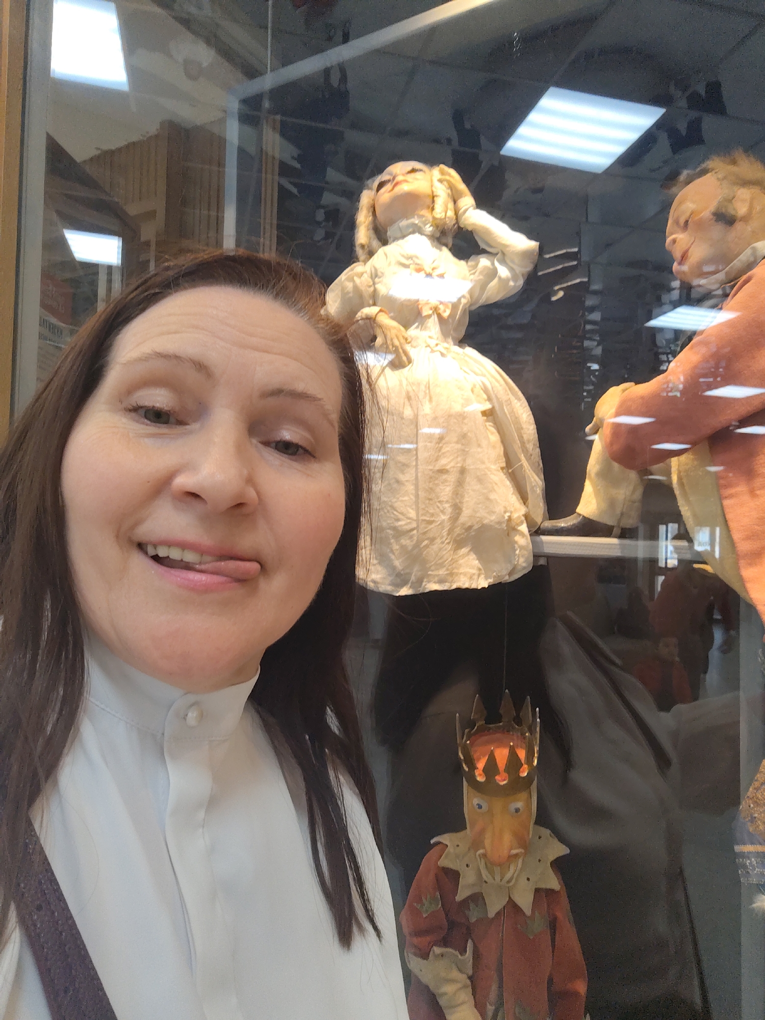 A person is taking a selfie in front of a display case of antique marionettes in Unknown, Unknown. The display includes a marionette of a woman in a cream-colored dress with her hand to her head, a marionette of a man in an orange coat, and a small marionette of a king with a crown. The person taking the selfie is sticking out their tongue.