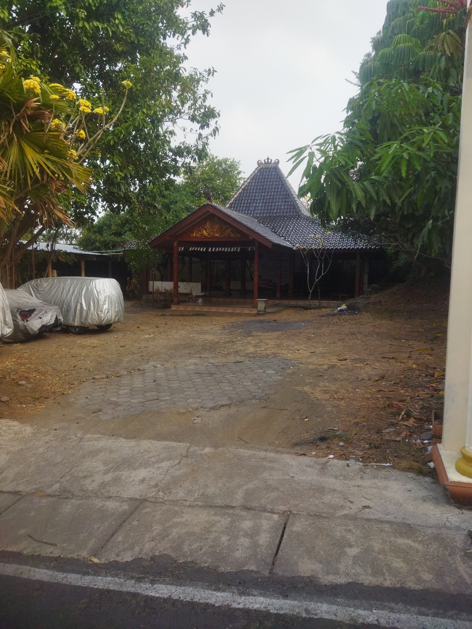 The image depicts an outdoor scene in the city of Wonogiri, Indonesia, featuring a traditional Javanese-style building with a multi-tiered, dark tiled roof. The building has an open-sided design with wooden pillars supporting the roof. In front of the building, there is a paved area with a patterned brick section and a concrete walkway. To the left, two vehicles are covered with silver tarps. The surroundings are characterized by dry, brown ground with scattered leaves and vegetation, including several trees with green foliage. The sky is overcast. The image is taken from a low angle, looking towards the building and its surroundings.