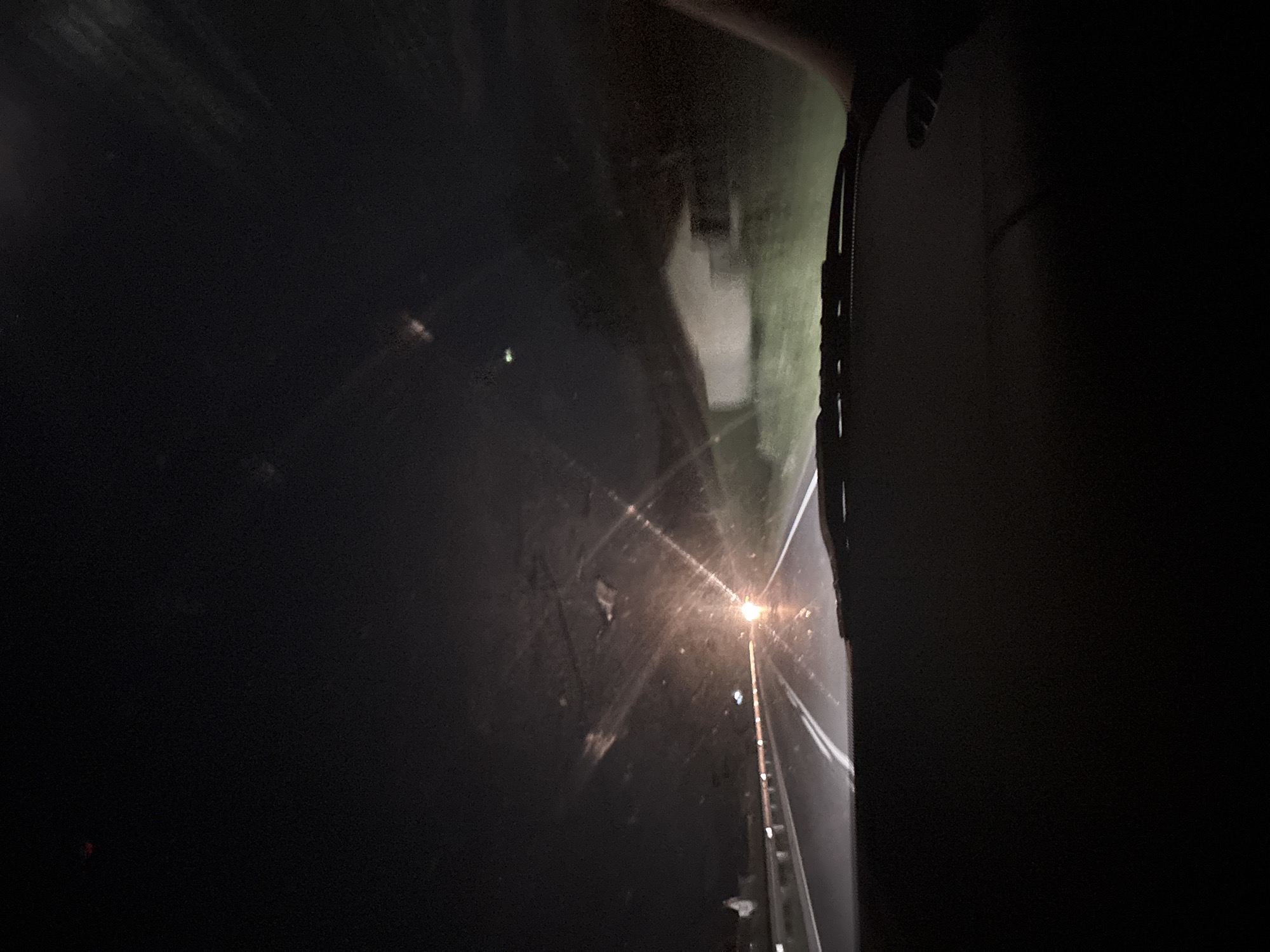 The image shows a night view of a road from inside a vehicle. The location is inferred to be near Koprivlen, Bulgaria, based on the prompt. A street light illuminates the road ahead, with its glare reflected on the wet surface. The roadside appears to be overgrown with vegetation. A guardrail runs along the right edge of the road. To the right of the center there is a dark object which seems to be some form of vehicle feature. Most of the image is obscured by darkness. No people are visible.