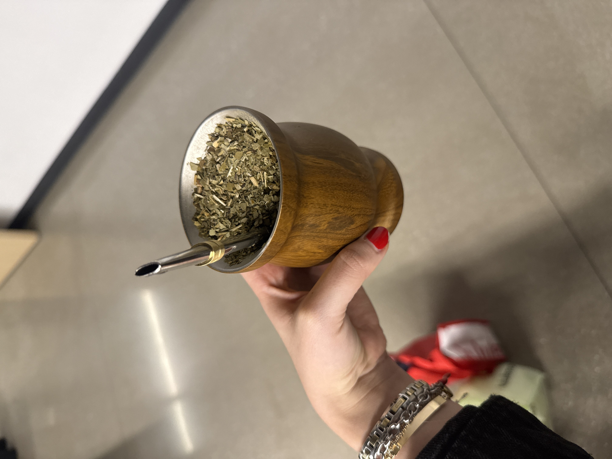 A close-up, high-angle shot captures a person's right hand holding a traditional mate gourd, prepared for consumption. The gourd is crafted from polished light-brown wood, featuring a shiny silver-toned metal rim. Inside, it's filled with dried yerba mate leaves and stems, characteristic of the popular South American beverage. A silver-colored metal bombilla, with a gold accent near its base, is inserted into the yerba mate, ready for drinking.
The hand shows red nail polish on the visible fingernails (thumb, index, and middle finger). The wrist is adorned with several metallic bracelets, one of which is a silver-toned watch with the brand name "GUESS" clearly visible on its band or clasp. A dark-colored fabric, likely a sleeve, is visible around the wrist.
The background is an out-of-focus indoor setting, featuring a light-colored, possibly reflective surface, which could be a countertop or table. Blurred diagonal lines and varied light and dark tones suggest other structural elements like walls or furniture in the soft-focus distance. The image provides no specific time of day or location cues beyond the given context of Mexico City, Mexico.