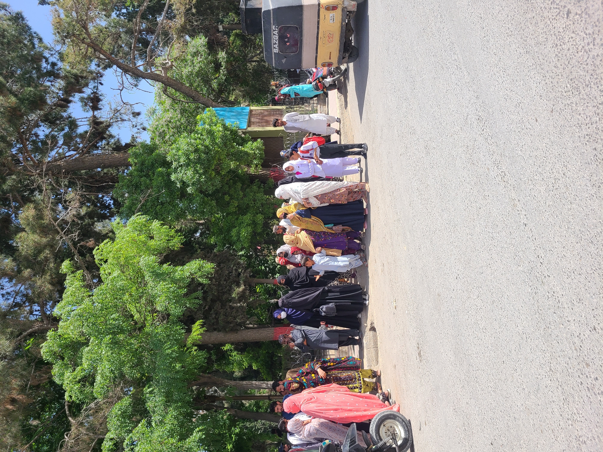 A long queue of people, primarily women and children, is seen standing on the side of a paved road in Quetta City Tehsil, Pakistan. The line extends from the foreground towards the background, beneath a canopy of green trees and under a clear blue sky, indicating daytime.
Many individuals in the queue are dressed in traditional clothing. The women wear a variety of colorful dresses, often with headscarves; notable colors include black, pink, purple, yellow, and white. Some children are visible in what appear to be white uniforms. A few men are also present in the line, wearing traditional tunics.
Along the road, several vehicles are visible. A yellow and black three-wheeled auto-rickshaw, or taxi, is prominent, with the word "SAZGAZ" clearly printed on its rear. Partially visible license plate numbers like "SAD-98" can be discerned. A motorcycle or scooter is parked in the foreground, and another red vehicle is partially visible further down the line.
The people appear to be patiently waiting in an orderly fashion. A light blue tarp or banner is visible near the front of the queue, possibly indicating a designated area or providing shade. The presence of numerous trees suggests a tree-lined street or a public park area. The scene depicts a common daily activity or gathering in a South Asian urban or semi-urban setting.