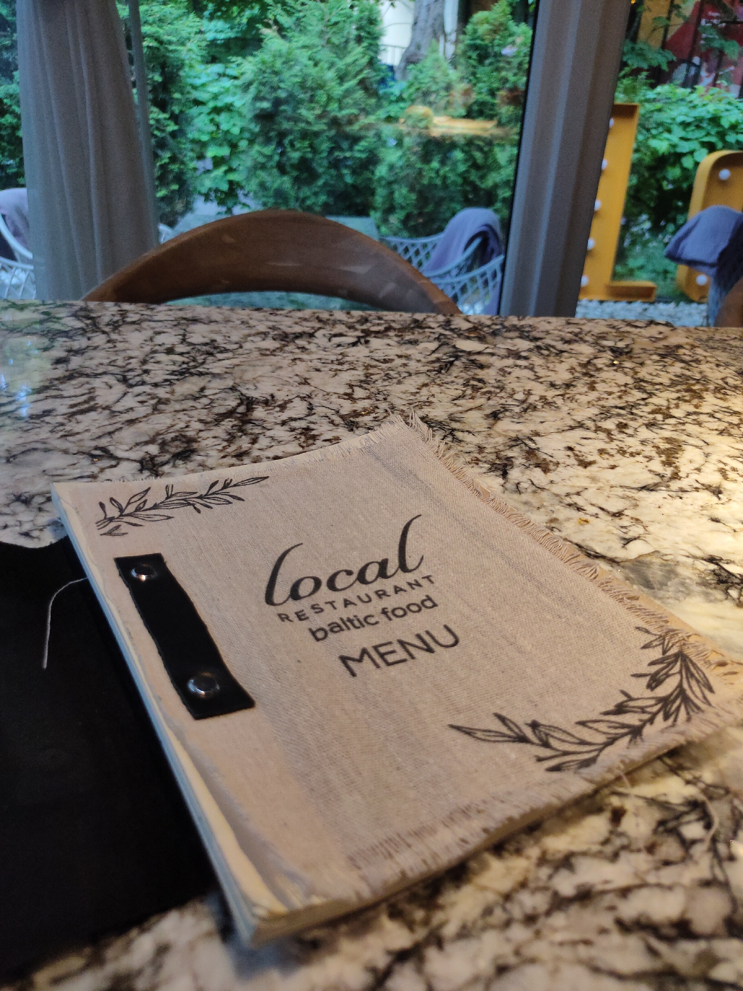 A menu is on a granite countertop in a restaurant with an outdoor seating area visible through a window. The menu is light brown with decorative green leaves printed on it. The text on the menu reads "local RESTAURANT baltic food MENU". A dark object, possibly a placemat or napkin, is tucked under the menu. In the background, through the window, lush green trees and outdoor seating with blue cushions are visible. Large illuminated letters spelling "LOVE" are also visible in the distance, positioned to the right. The overall ambiance suggests a casual dining experience in a garden setting, likely in Svetlogorsk, Russia, a resort town known for its coastal location and German architectural influences, which could explain the prominent "LOVE" sign.