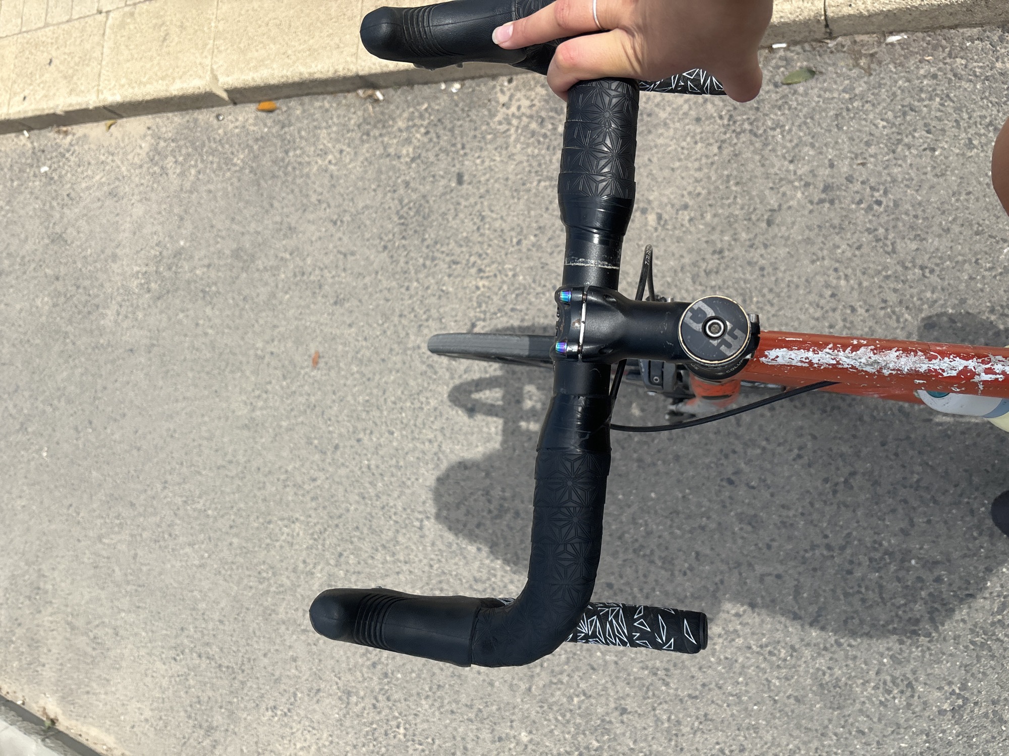 An overhead shot captures a person's perspective of riding a bicycle on a paved road next to a concrete curb under bright sunlight, likely in Tel-Aviv, Israel. The strong shadows suggest a clear, sunny daytime.
The primary subjects are the bicycle's handlebars and stem, along with a person's hand gripping the right side of the handlebars. The person's right hand, adorned with a simple ring on the index finger, holds the black handlebar tape. A small portion of the rider's leg or thigh is visible on the far right.
The bicycle features black drop handlebars, mostly wrapped in black tape with a repeating geometric, star-like pattern. Towards the drops, the tape has a distinctive white geometric design against a black background. Black brake/shifter hoods are visible at the bends of the handlebars. The black stem connecting the handlebars to the frame is secured with iridescent, rainbow-colored bolts. The top cap of the headset is black with a silver trim and clearly displays the text "COLUMBUS" and "PFP." below it.
A section of the bicycle's frame, visible below the stem, is orange or red with significant white scuffing or paint distress. A sliver of the front wheel's tire is also visible. The ground is a light gray, coarse paved surface (asphalt or concrete), with a distinct concrete curb running along the left edge of the image. Small bits of debris are scattered on the pavement near the curb.