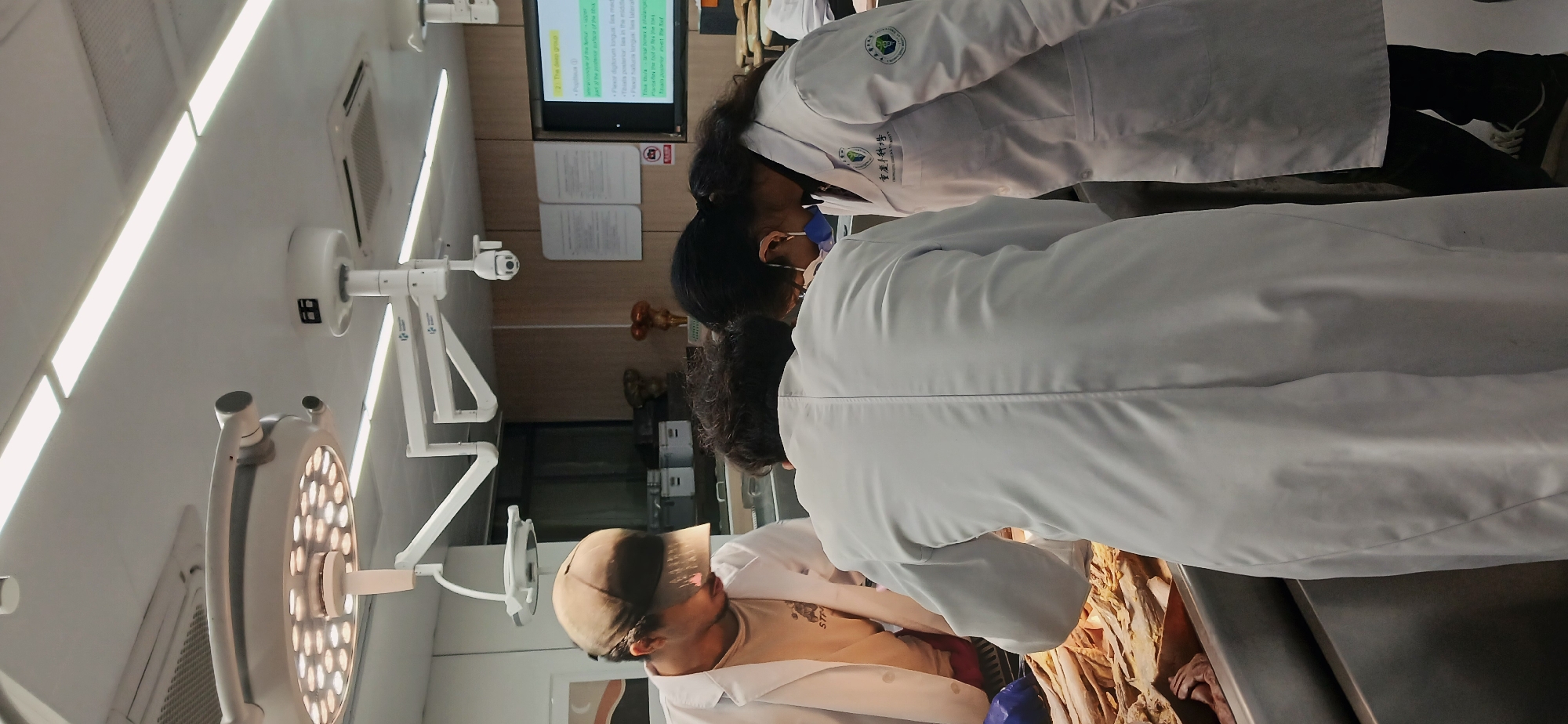 In a bright, sterile environment reminiscent of a laboratory or operating room, a group of individuals in white lab coats are gathered around what appears to be a dissection table. The setting includes surgical lighting and a screen displaying medical information, suggestive of a scientific or medical context. A close examination of the lab coats reveals the logo of Chongqing Medical University and the words "重庆医科大学", indicating the location is likely in Chongqing High-tech Zone, China. The focus of the group's attention is directed towards the contents of the dissection table, where anatomical samples or a cadaver are likely present. The participants are engaged in what seems to be an educational activity, potentially involving medical students or researchers studying anatomy.