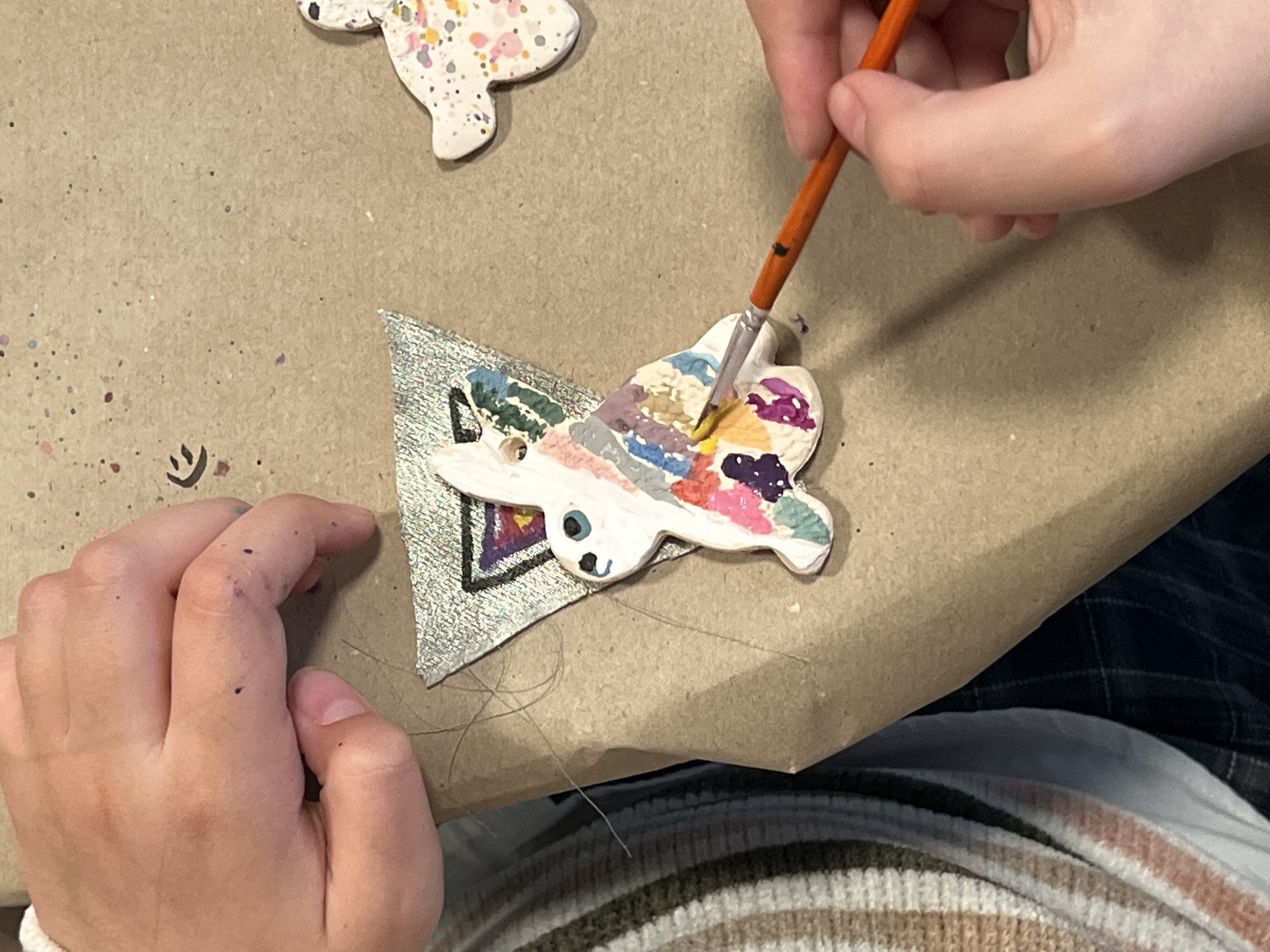 A person is painting a small, ceramic, animal-shaped ornament with a paint brush. The ornament is mostly white, with sections of various colors including pink, blue, green, purple, and yellow. A painted silver triangle is underneath the ceramic ornament. The person's hands are visible; one hand holds the paintbrush while the other rests near the ornament. There are purple spots of paint on the person's left hand. Another similar ceramic ornament, with a white base and multicolored splatters, sits in the top left corner of the frame. The surface beneath the objects is a brown, slightly textured material. The person is wearing a sweater with white, brown and green stripes. The location and city of Belgrade, Serbia are unknown.